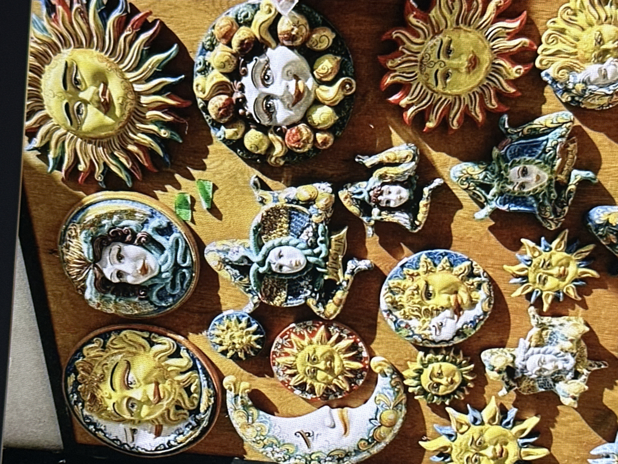 The image displays a collection of ceramic decorative items, predominantly featuring sun and moon motifs, as well as stylized faces. These objects appear to be displayed on a wooden surface. In the upper left quadrant, a circular ceramic piece depicts a sun with a human face, radiating yellow rays. Adjacent to it, a similar circular piece shows a woman's face with elaborate, blue and green hair. Another large, circular sun face with colorful rays dominates the upper right.
Below these, a crescent moon with a human face is visible on the left, adorned with floral patterns. To its right are smaller circular sun faces. Several other pieces feature stylized faces, some with intricate, serpentine hair like Medusa, and others depicting sun motifs. The overall aesthetic suggests a theme of celestial bodies and classical or mythological figures, likely crafted in a ceramic artisan tradition. There are no people or actions depicted in the scene, and the environment is a static display of these decorative objects.