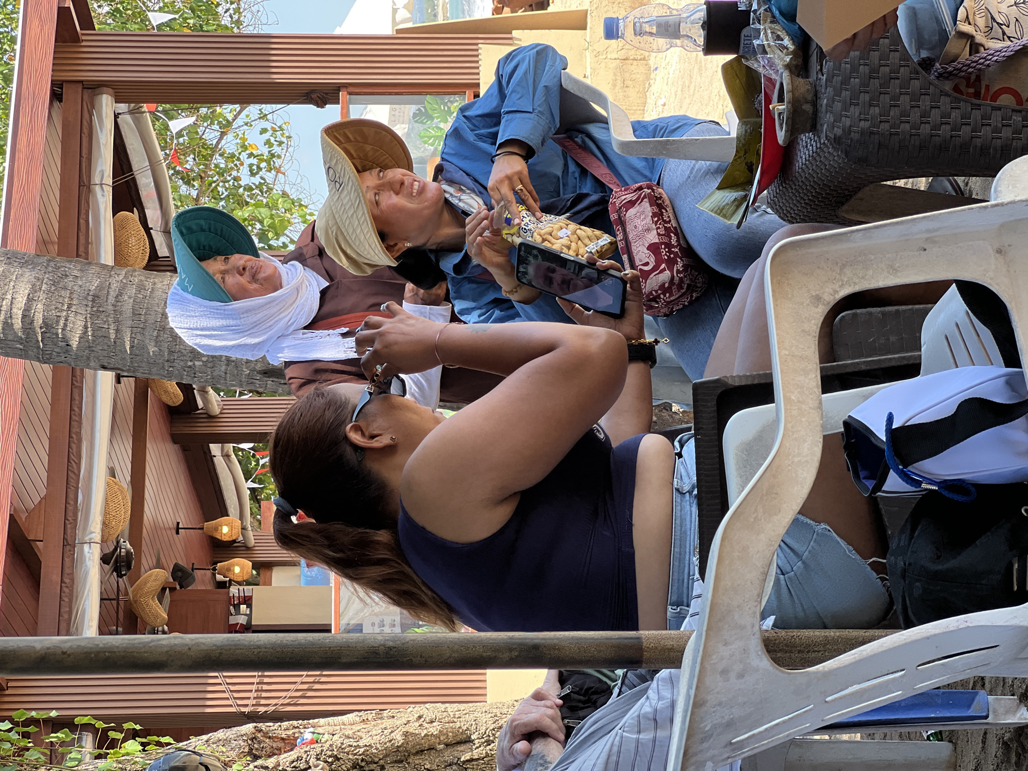In the city of Kuta, Indonesia, three individuals are seated at a table outdoors, likely at a beachfront restaurant or cafe, as suggested by the palm tree, sand on the ground, and a glimpse of the ocean in the background. The focus is on the interactions between two women in the foreground. One woman, wearing a dark blue tank top and denim shorts, is looking down at a phone held by another woman. This second woman, wearing a blue collared shirt and a wide-brimmed straw hat, is holding a bag of peanuts and also looking at the phone. A third woman, further back and partially obscured, wears a white headscarf and a brown garment, also looking towards the phone. The table is laden with food items, including what appears to be food wrapped in banana leaves and a bottle of water. The scene is lit by natural sunlight, casting shadows and highlighting the casual, relaxed atmosphere of a tropical setting.