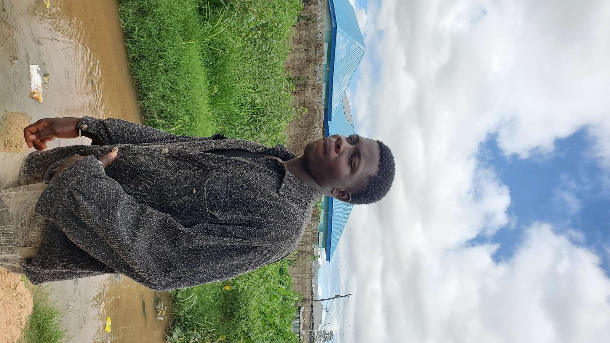 In Jalingo, Nigeria, under a sky with scattered white clouds against a blue background, a young Black man stands outdoors. He is viewed from the chest up, facing towards the right of the frame, with his head turned slightly towards the viewer. He has dark, short, curly hair and is wearing a dark, textured, button-up shirt over a dark t-shirt, and light-colored trousers. His left hand is inside the front pocket of his trousers. Behind him, a low stone wall is visible, topped with barbed wire in some sections, separating the foreground from the background. Beyond the wall, there are two buildings with distinctive bright blue, pitched roofs. Lush green vegetation, including tall grasses, grows in front of and behind the wall. In the lower foreground, a shallow body of brown, muddy water is present, with some debris, including a white and orange package, visible on its surface and along the bank.