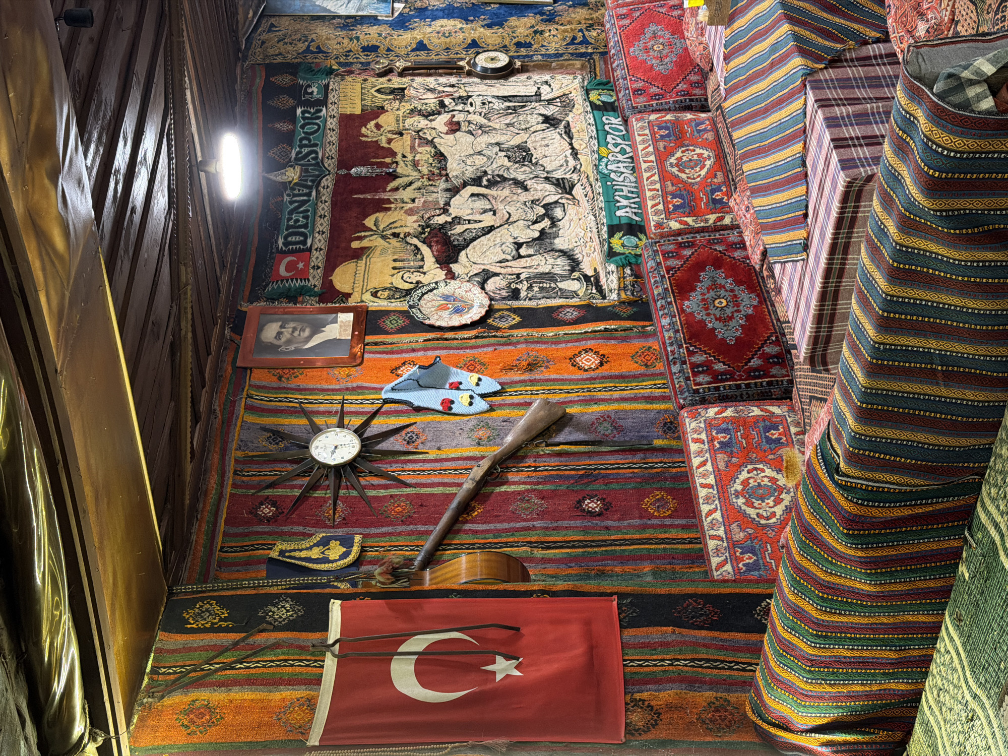 The image depicts an interior space in Pamukkale, Turkey, characterized by a dense collection of textiles and cultural artifacts covering the walls. A Turkish flag is prominently displayed near the bottom, positioned above the image. Above it is a stringed instrument, partially obscured by a long, brown, wooden object that resembles a rifle. A starburst clock is mounted on the wall next to a pair of blue slippers. A framed portrait hangs on the left. Banners, carpets, and other patterned fabrics create a visually rich backdrop. A bright light source illuminates the scene from the upper left. The walls appear to be made of wood paneling.