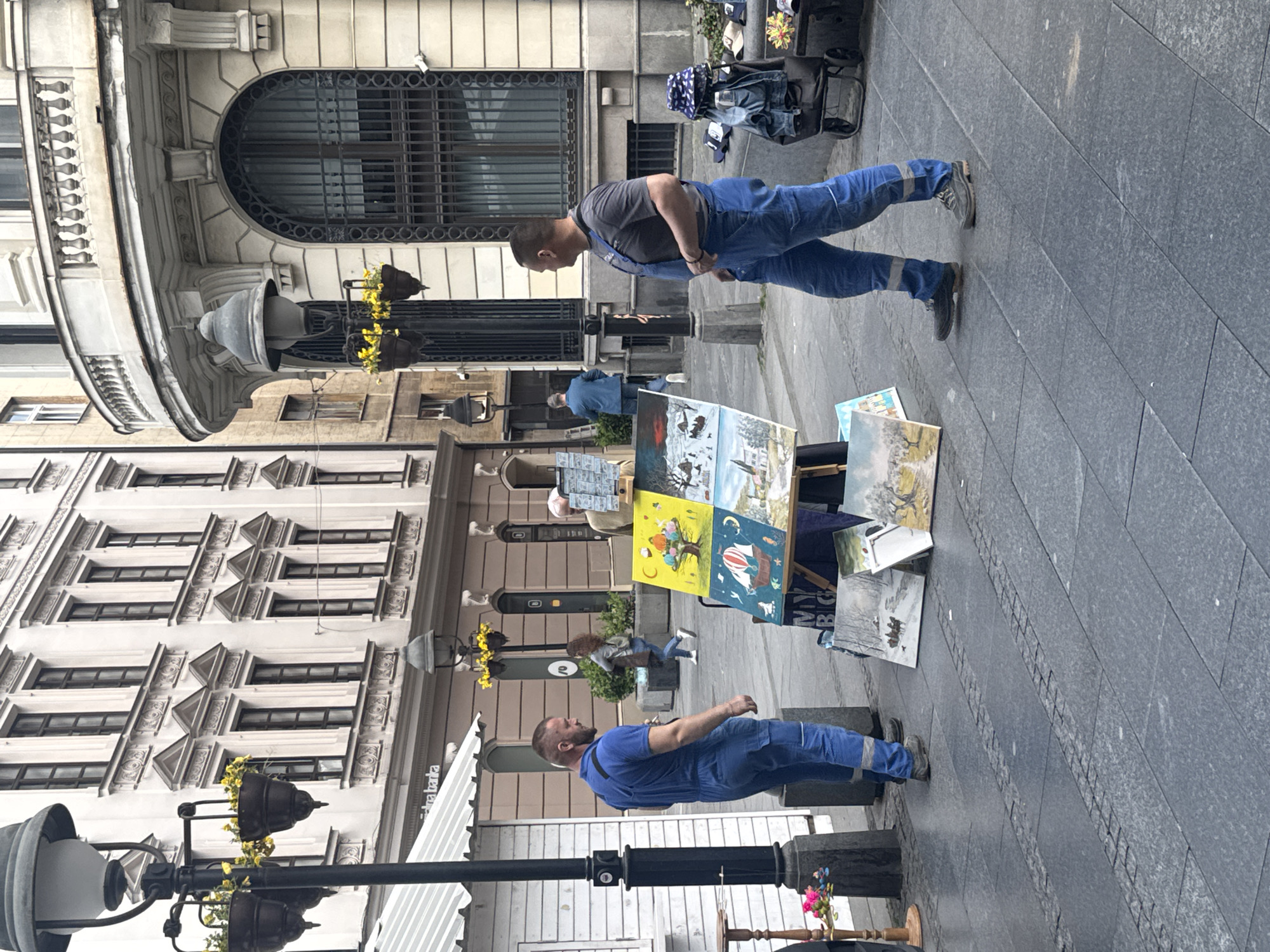 This image captures a vibrant street scene in Belgrade, Serbia, set on a clear day. The view is dominated by ornate, multi-story stone buildings with classical architectural features, including arched windows with intricate grilles and decorative balconies.
In the foreground and midground, two men in blue work overalls stand on a dark grey tiled pavement. The man in the midground, wearing a grey t-shirt, stands facing left with his hands clasped. Closer to the viewer, another man with a beard, also in blue overalls and a black t-shirt, stands in a similar posture.
Between these men, a street vendor's display is set up, showcasing various artworks. These include landscape paintings, some depicting winter scenes, and more colorful, whimsical illustrations on a cube-shaped stand featuring images of hot air balloons, animals, and fantastical elements. Partially visible text on one canvas reads "MY BC". Another small display with hats is visible further back.
Several other people are scattered throughout the background, some walking, and a woman is seated on a bench. The street is adorned with elegant black streetlights featuring hanging baskets filled with yellow flowers. A white awning on the left side prominently displays the word "banka." The scene depicts a lively pedestrian zone in an urban environment.