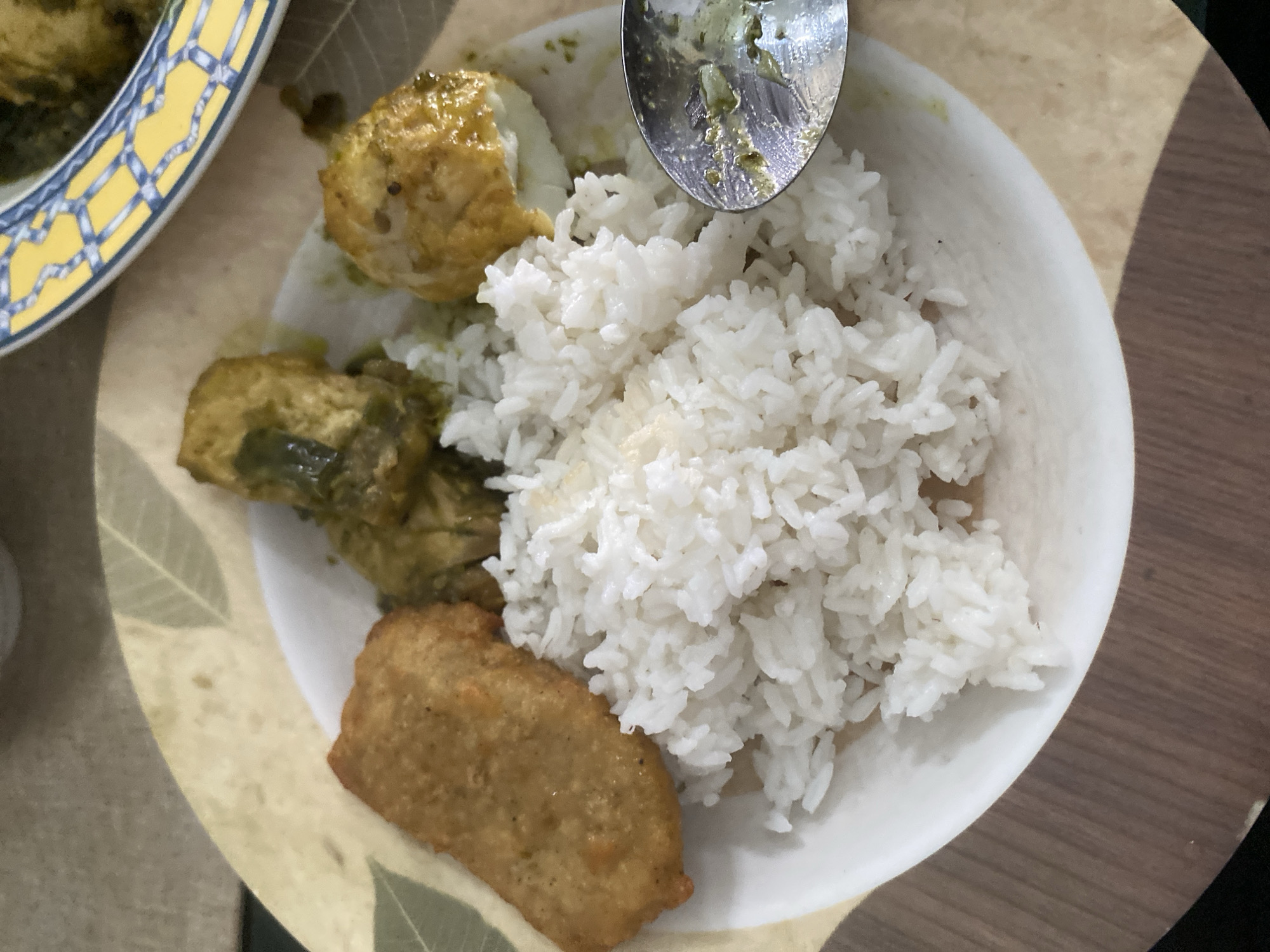 A plate of food is shown from a top-down perspective. The plate contains white rice, a piece of fried food that appears to be a fritter or croquette, two pieces of chicken or tofu in a green curry sauce, and a hard-boiled egg cut in half and also coated in sauce. A spoon with remnants of green sauce is positioned on the right side of the plate. The plate rests on a placemat with a leaf pattern. The background is a wooden table. This scene is located in Dukuh, Indonesia.