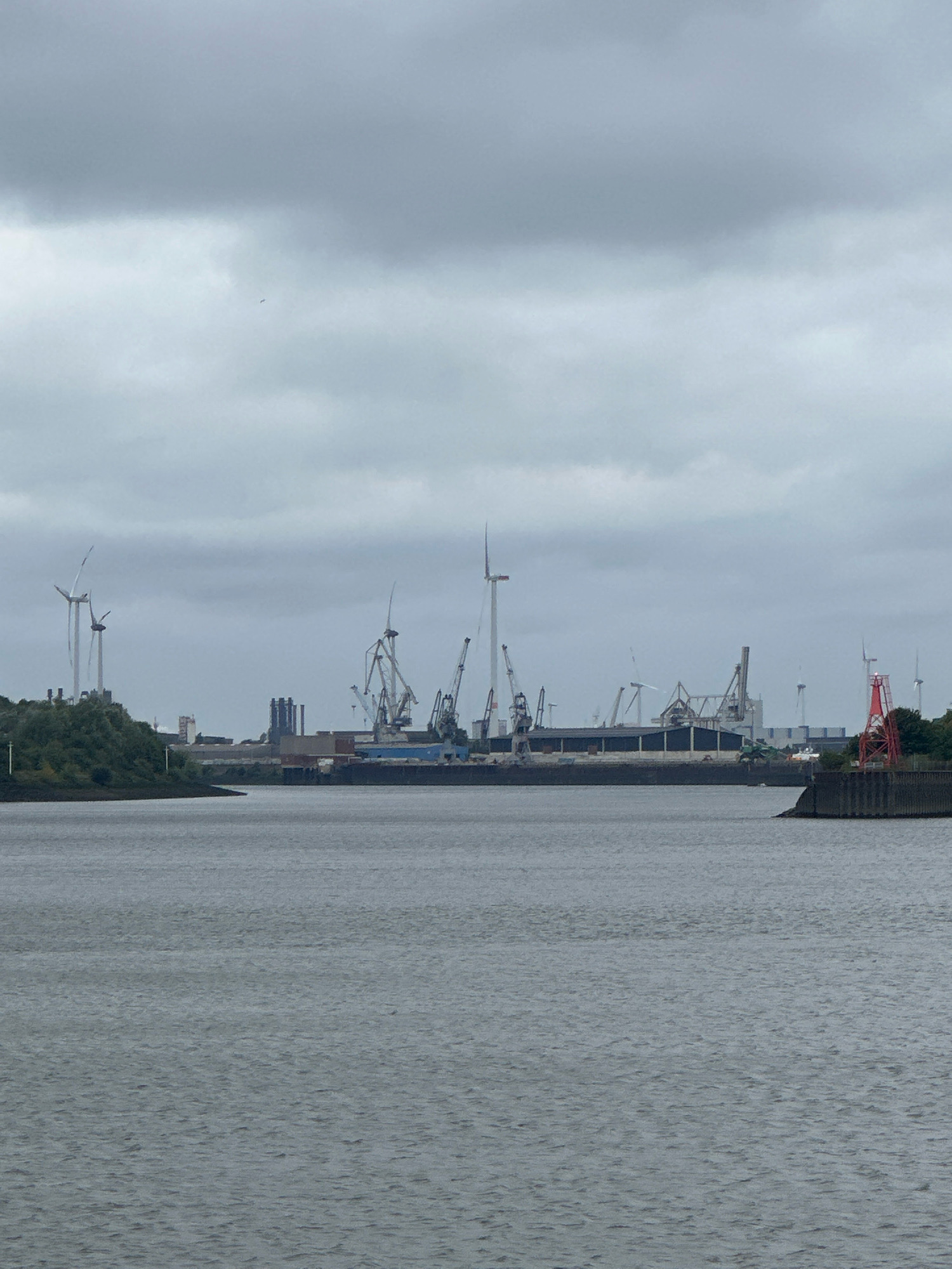 The image depicts a cloudy day over a harbor or industrial waterfront. In the foreground, a body of grey water stretches across the bottom of the frame. In the middle ground, the harbor features a long, dark industrial building with a blue roof. Several tall cranes, of varying designs, are positioned along the waterfront, some appearing to be in use. Wind turbines are visible on a green, hilly area to the left of the frame. Further in the distance, more industrial structures and cranes are present. A red, lattice-like structure with a light on top stands on the right side, marking a point in the water. The sky is overcast with grey clouds. There are no people visible in the image. The scene suggests an active industrial or port area.
