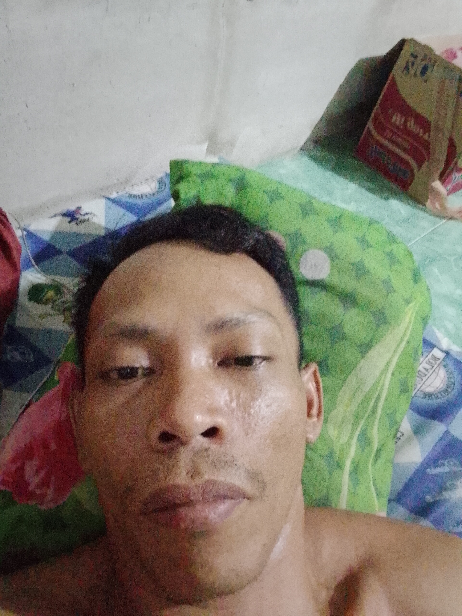 A shirtless, adult male is lying on a bed with a blue and white patterned sheet and a green patterned pillow. There is a red and brown cardboard box near his head. The photograph appears to be taken in a dimly lit room, possibly in Bukit Ketok, Indonesia, based on the prompt. The man is looking directly at the camera.