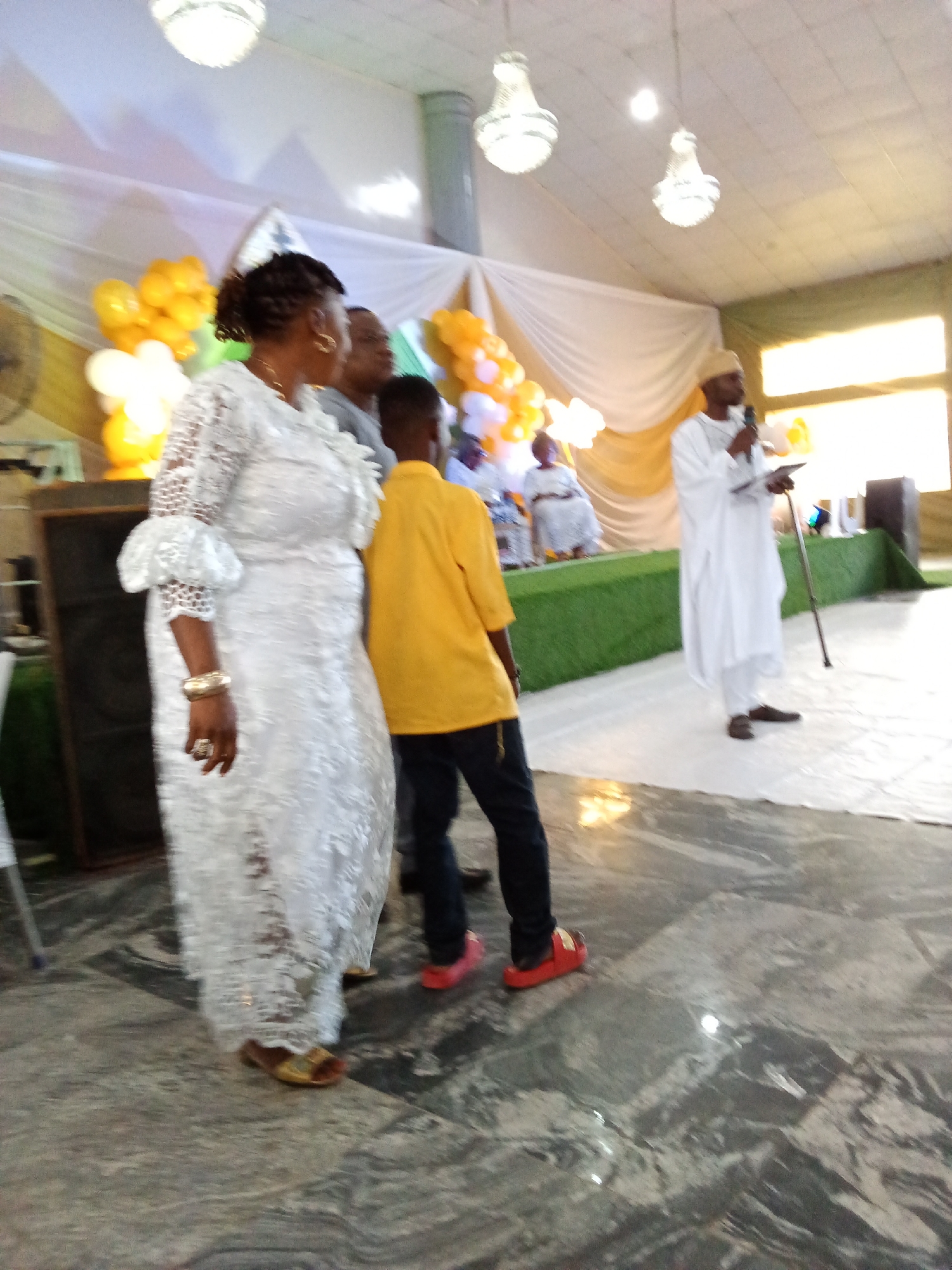 In Barden Yana, Nigeria, at what appears to be a celebratory gathering, several individuals are visible indoors. The scene is decorated with draped white and yellow fabric and illuminated by chandeliers. A woman in a white lace dress stands in the foreground, facing left, with her arm bent and hand resting on her hip. To her right, a young boy in a yellow shirt and dark pants is seen from the back. Further in the background, a man in a white robe and cap speaks into a microphone while holding a tablet and a walking stick. Two other figures are seated on a raised platform behind him. The floor is a reflective, marbled surface.