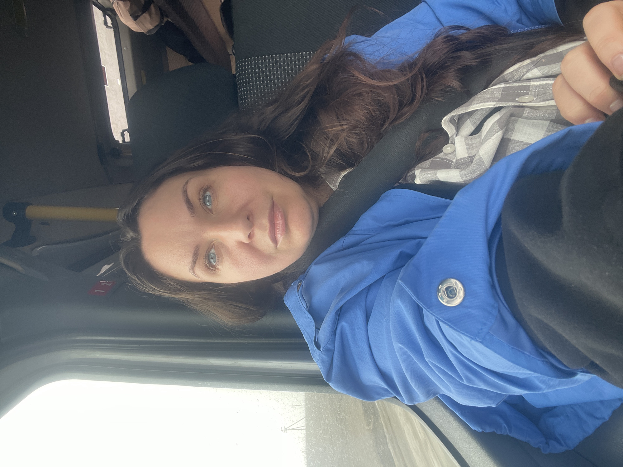 A young woman with brown hair and blue eyes is wearing a blue jacket and a gray plaid shirt. She is in the passenger seat of a vehicle, wearing a seatbelt. The vehicle appears to be in Bol'shoi Mashlyak, Russia. The outside environment visible through the window appears bright and overcast.