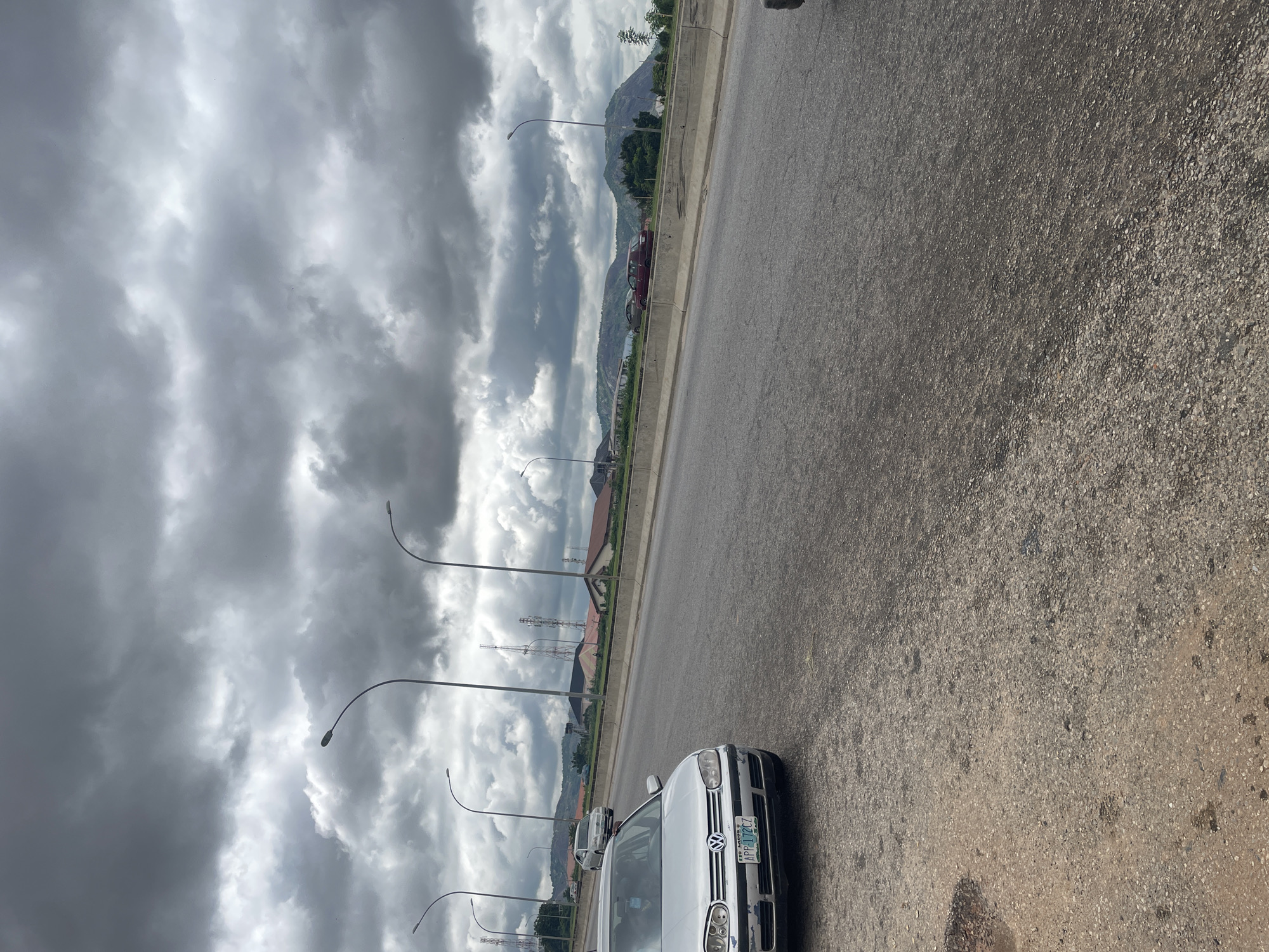 The image depicts a cloudy day on a highway in Kubwa, Nigeria. A silver Volkswagen Golf is parked on the shoulder of the road, its front bumper showing some wear and tear. The license plate reads "APP 112 CZ". Further down the road, other vehicles are visible, including a silver sedan and a maroon car. In the background, residential buildings and communication towers rise against a backdrop of hills and mountains under a heavy, overcast sky. The road surface is asphalt with visible gravel and some dark patches.