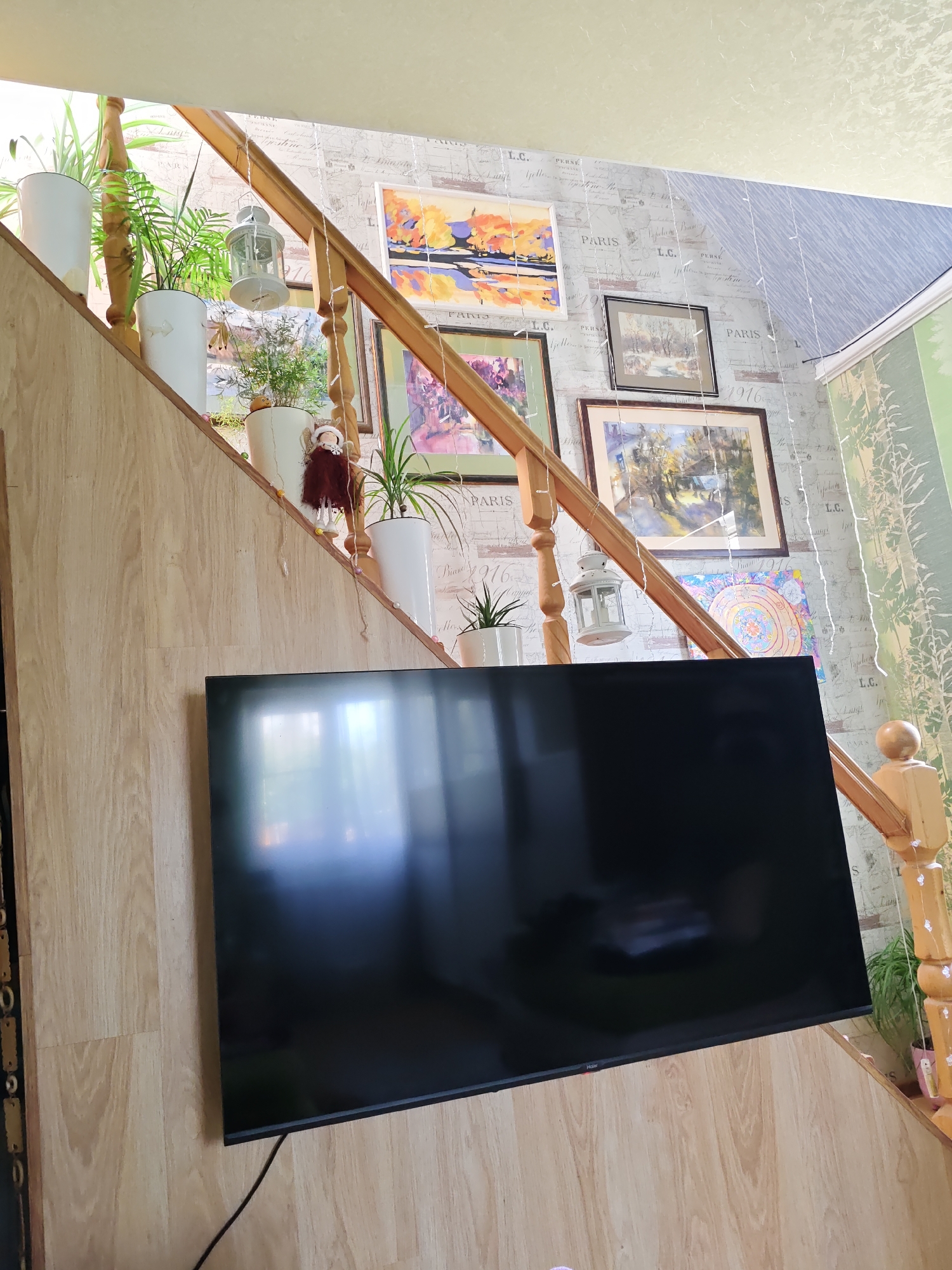 The image shows a television screen mounted on a wooden paneled wall next to a staircase. The staircase has a wooden banister and the steps are also paneled in wood. Several potted plants are placed on the steps, along with decorative lanterns and small hanging ornaments. The wall behind the staircase is decorated with wallpaper featuring a pattern of text and drawings, and several framed pictures are hung on it. The overall scene appears to be an interior residential space, possibly a hallway or living area with a staircase. The lighting suggests it is daytime, with natural light coming from above. No people are visible in the image. The location in the city of Rostov-on-Don, Russia is not discernible from the visual information provided in the image.