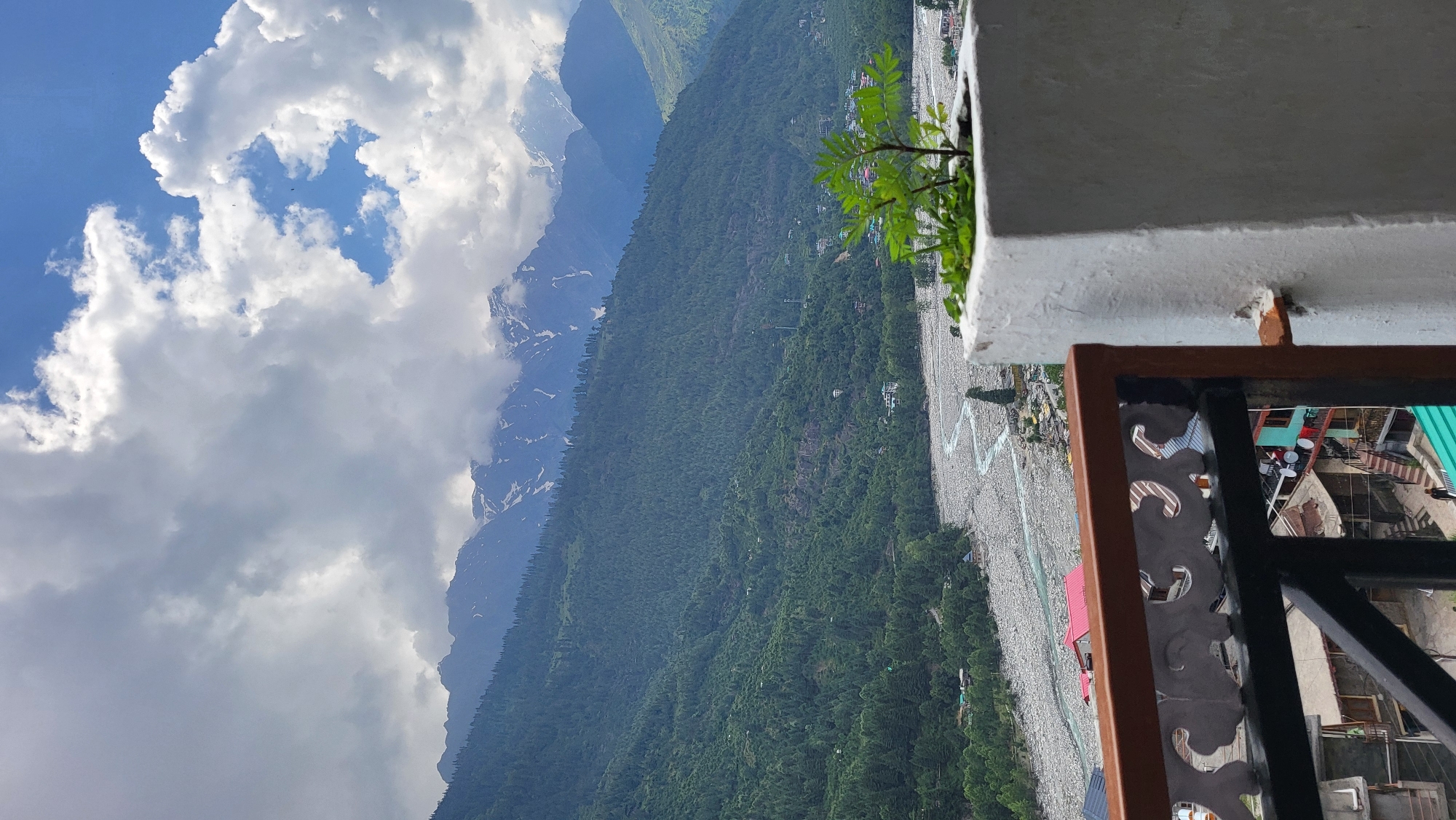 This image captures a panoramic view of a mountainous valley, observed from a balcony, likely in Vashisht, India, given the regional characteristics.
In the immediate foreground, to the bottom left, a dark brown and black metal balcony railing with white, ornate carvings is visible. To the bottom right, a white, rough-textured concrete planter holds a small green plant with serrated leaves.
The midground reveals a deeply carved valley. A wide riverbed, predominantly gray with numerous smooth, rounded rocks and boulders, meanders from the upper left towards the lower right. A narrow, light blue-green stream of water flows through this rocky bed. Along the riverbanks and extending up the lower mountain slopes are dense evergreen forests, predominantly dark green, indicating coniferous trees. Scattered throughout these forested slopes and along the river are small clusters of buildings with multi-colored roofs (red, green, blue, brown), characteristic of a village or town. Some buildings exhibit multiple satellite dishes. One human figure in dark clothing is discernible walking on a path in the lower left section of the midground.
In the background, towering mountains dominate the horizon. The lower and mid-sections of these mountains are heavily forested with dark green trees. Higher elevations, particularly on the right side of the frame, feature snow-capped peaks with visible patches of white snow against dark rock faces. The sky above is partially overcast with large, fluffy white and gray clouds, interspersed with clear patches of vibrant blue sky. Two small, dark specks, possibly birds, are visible against the blue sky in the upper right quadrant.