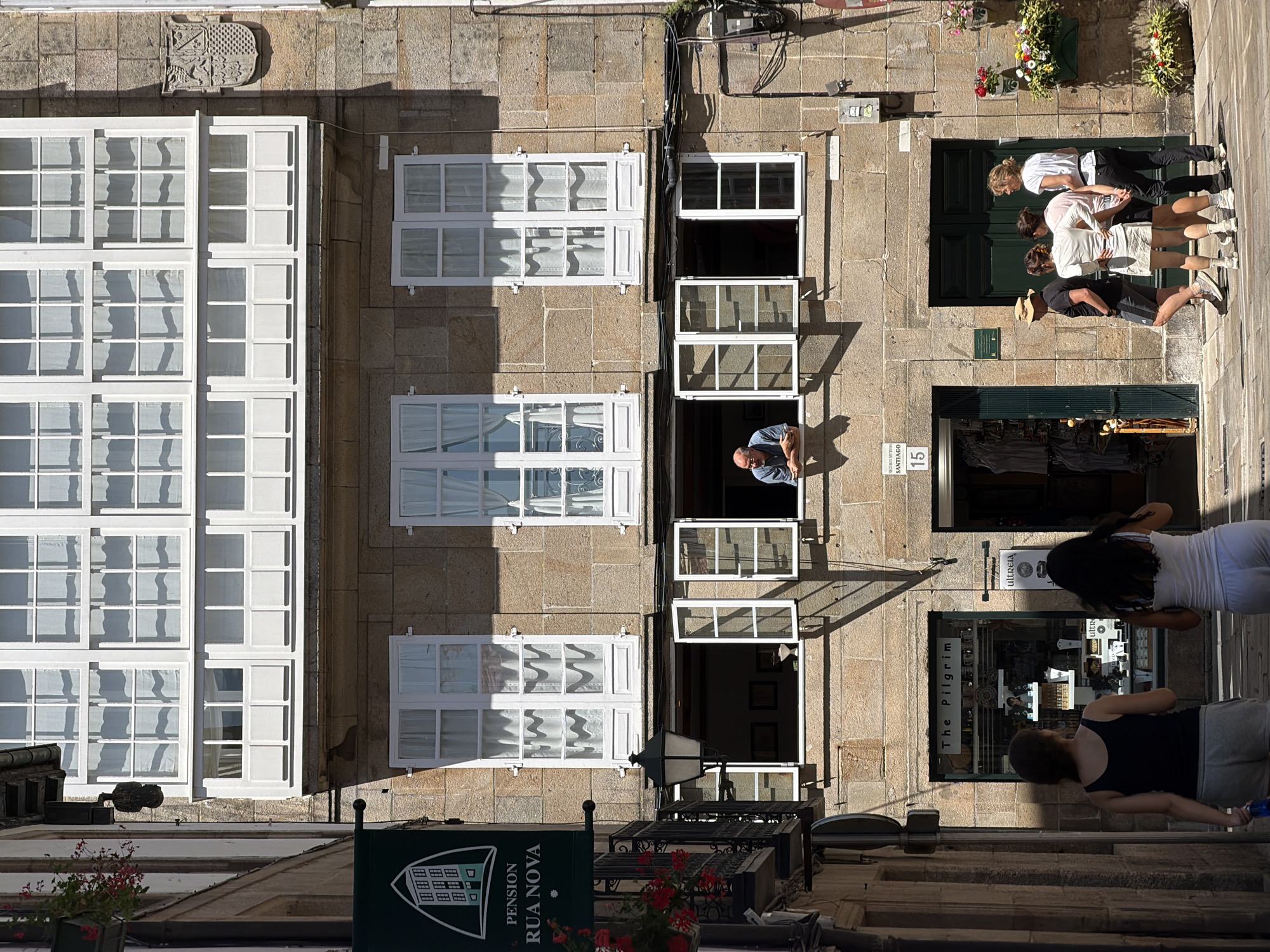 This is a ground-level exterior shot of a stone building in Santiago de Compostela, Spain, featuring a residential facade above shopfronts. The building has multiple levels of windows with white frames and shutters. On the first floor of the shops, there is a green awning for "PENSION RUA NOVA." Below that, a balcony with red flowers overlooks the street. Further down, a shop window reads "The Pilgrim." A man is visible leaning out of a second-story window, looking downwards. Four individuals stand near the entrance of another shop on the ground floor, appearing to be a group of tourists. The scene is illuminated by bright sunlight, casting strong shadows.