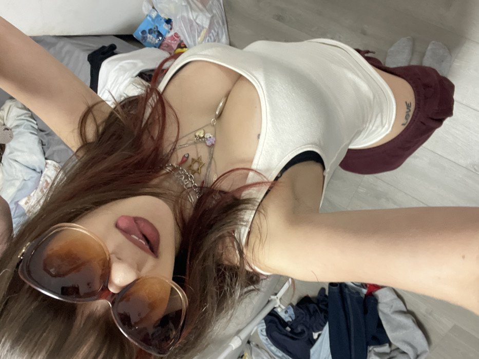A selfie captures a young woman lying down. She's wearing large, gradient sunglasses and sticking out her tongue slightly. Her hair is reddish-brown, and she's adorned with layered necklaces. She is wearing a white tank top and maroon sweatpants. The background is cluttered with laundry and other items, giving a casual, intimate feel to the image. The photograph is taken from a high angle, emphasizing her face and chest.