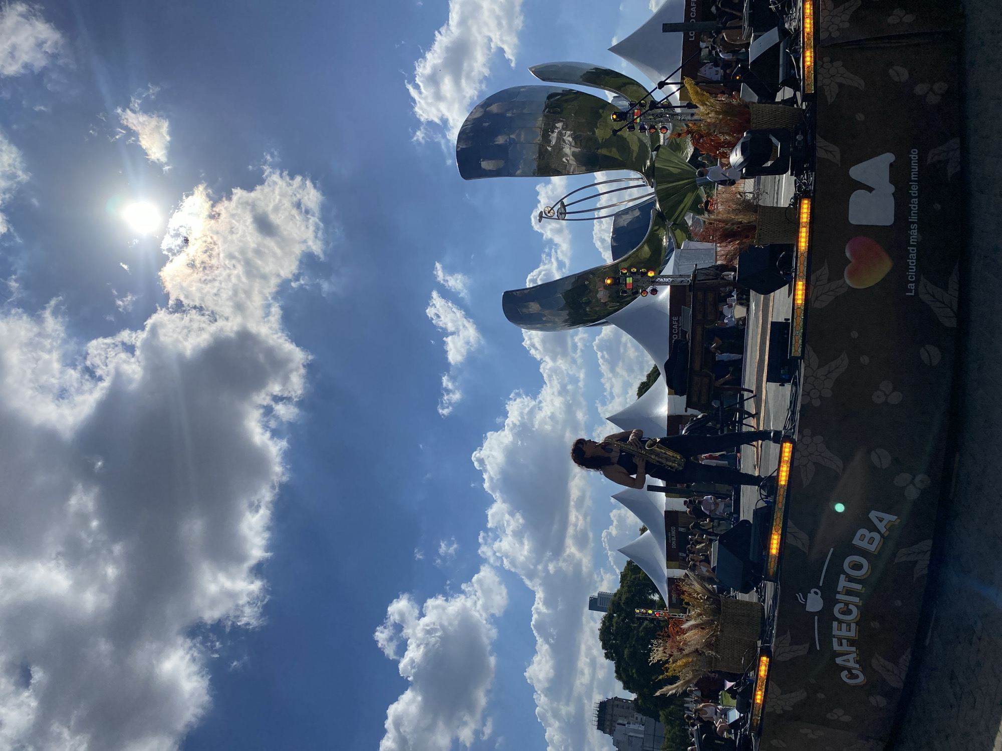 The image depicts a vibrant outdoor event in Buenos Aires, Argentina, under a bright, partly cloudy sky. The sun, partially obscured by clouds, casts a strong light over the scene.
In the foreground, a person with reddish-brown hair is actively playing a saxophone on a performance stage. They are dressed in dark attire. The stage is adorned with baskets filled with dried grasses or reeds and features a strip of bright yellow lights along its front edge.
Dominating the mid-ground is the Floralis Genérica, a large, gleaming metallic flower sculpture, reflecting the blue sky and clouds. Several white tents or marquees are set up around the stage area, suggesting an event with multiple vendors or activity zones. Beyond these, trees and urban buildings are visible in the distance, and glimpses of people in the background indicate an audience or attendees for the event.
Prominently displayed on the side of the stage are various texts: "CAFECITO BA" accompanied by a coffee cup icon, and the stylized letters "BA" next to a heart, underneath which reads "La ciudad más linda del mundo" (The most beautiful city in the world). Other readable texts include "DON BLANCO" and "LO CAFÉ." The combination of the iconic Floralis Genérica sculpture and the "BA" branding firmly places the setting in Buenos Aires, highlighting a lively public gathering or festival, possibly centered around coffee, given the "Cafecito BA" text. The bright daylight suggests it is daytime, with scattered cumulus clouds adding texture to the blue sky.