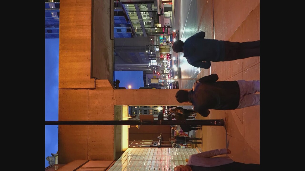 The image depicts a street scene in Vancouver, Canada, at dusk. The perspective is from a pedestrian walkway, looking out onto a street with vehicular traffic. Two men are walking away from the camera on the walkway, one in a dark leather jacket and light-colored pants, the other in a dark hooded sweatshirt and dark pants. Other pedestrians are visible in the background on the walkway. The street is lined with buildings and streetlights. The sky above is a deep blue, indicating twilight. The architecture suggests an urban environment with commercial buildings.