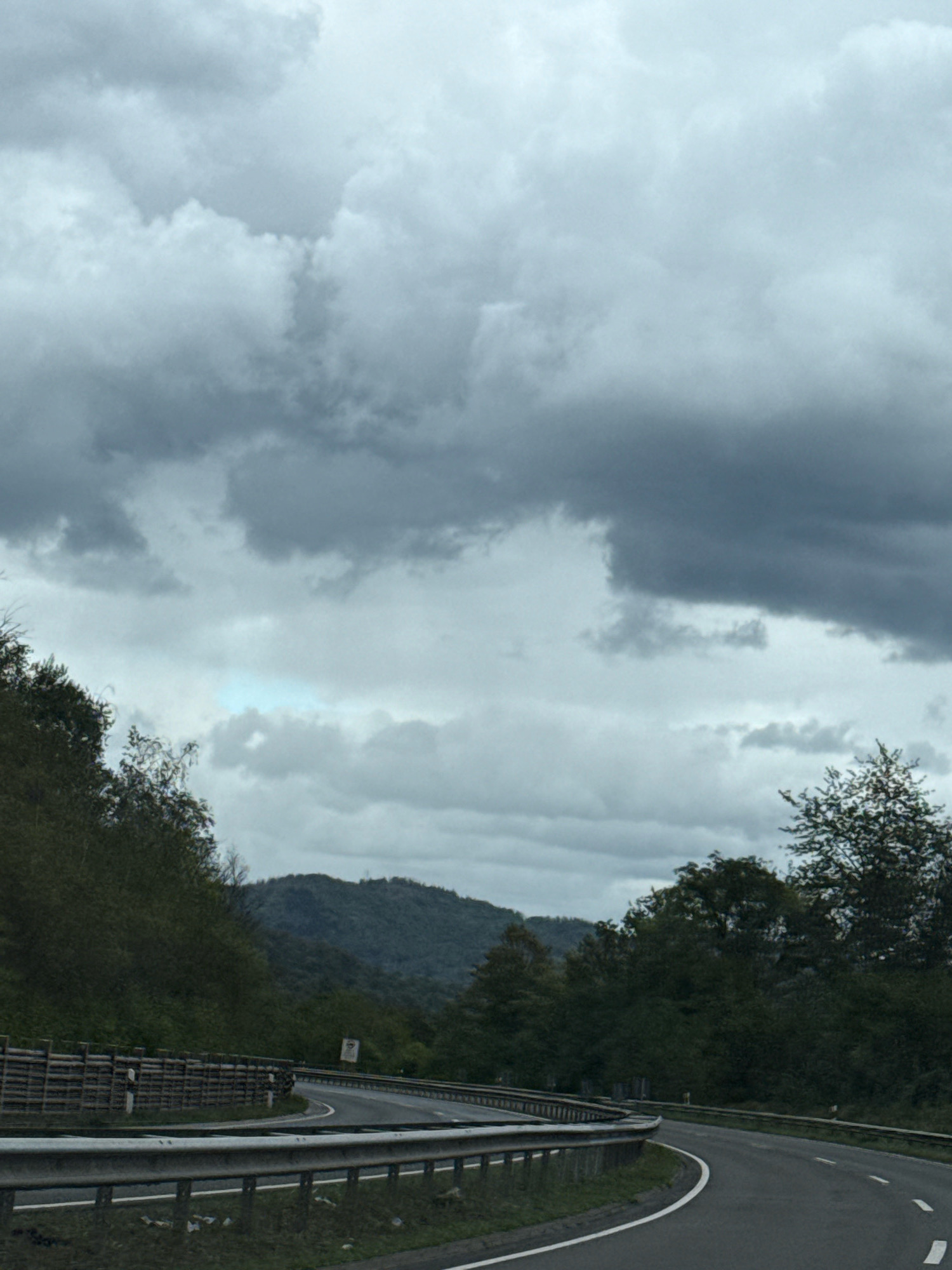 The image depicts a scenic view of a winding road in a rural, mountainous area, likely a highway or a major road, with guardrails visible on both sides. The road curves to the right in the foreground, leading towards a dense forest and rolling hills in the background. The sky is heavily clouded with dark, dramatic formations, suggesting an overcast or approaching storm. Vegetation, including trees and bushes, lines the sides of the road and covers the hills. The overall atmosphere is somber and natural, with no visible people or vehicles. The scene does not contain any elements that would specifically identify it as being located in Herzberg, Germany.