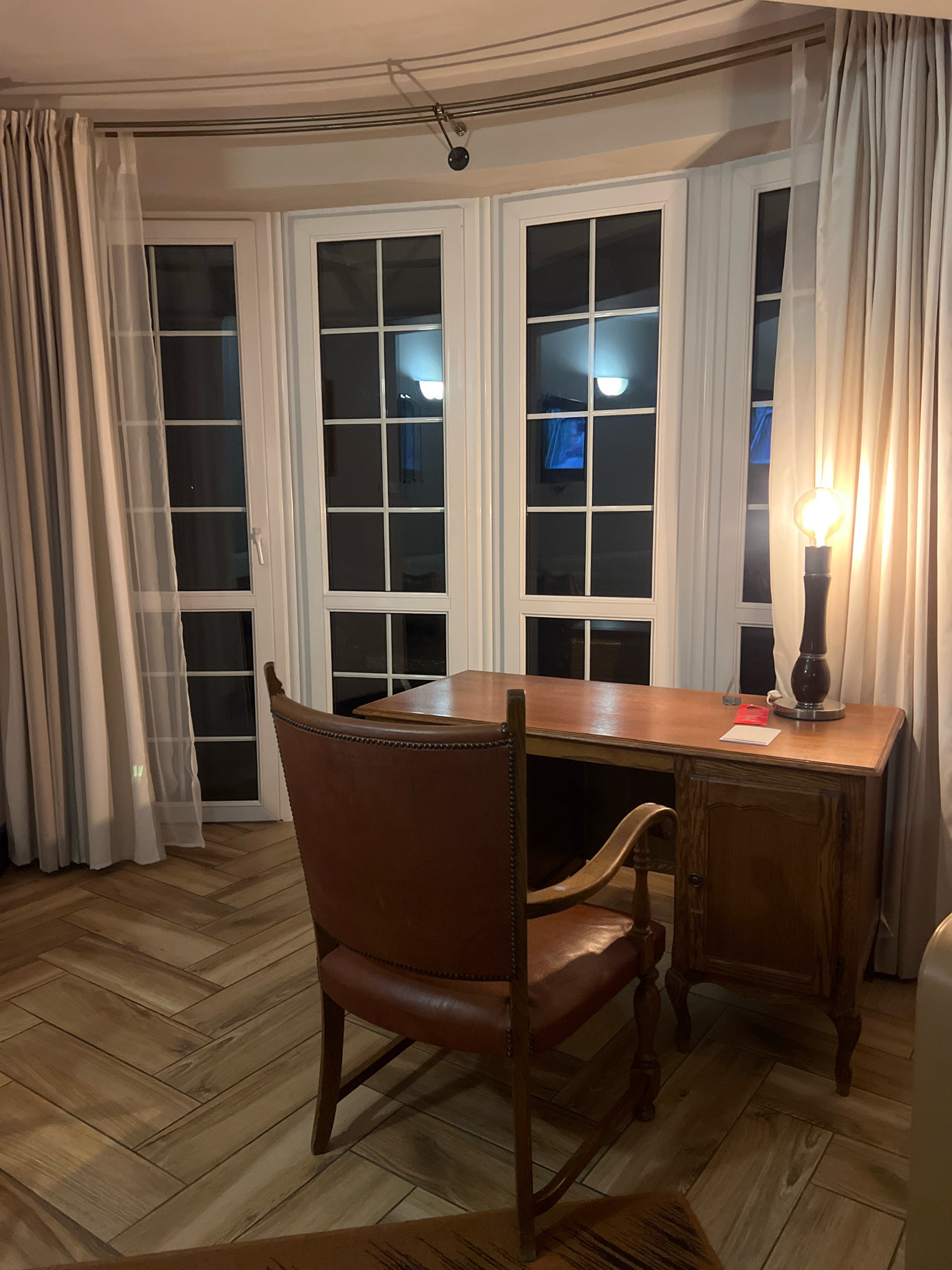 The image depicts an interior scene, likely a room in Mrągowo, Poland, furnished with a desk and chair. The room features large, multi-paned windows with white frames, covered by beige curtains. A wooden desk with a single drawer and a cabinet is positioned in front of the windows. On the desk, a vintage-style lamp with a bright bulb illuminates the space. A brown leather armchair with a wooden frame is placed in front of the desk. The floor is covered with light-brown wooden planks arranged in a herringbone pattern. The scene is illuminated by the desk lamp, with some light reflecting from the windows, suggesting it is nighttime or a dimly lit indoor space.