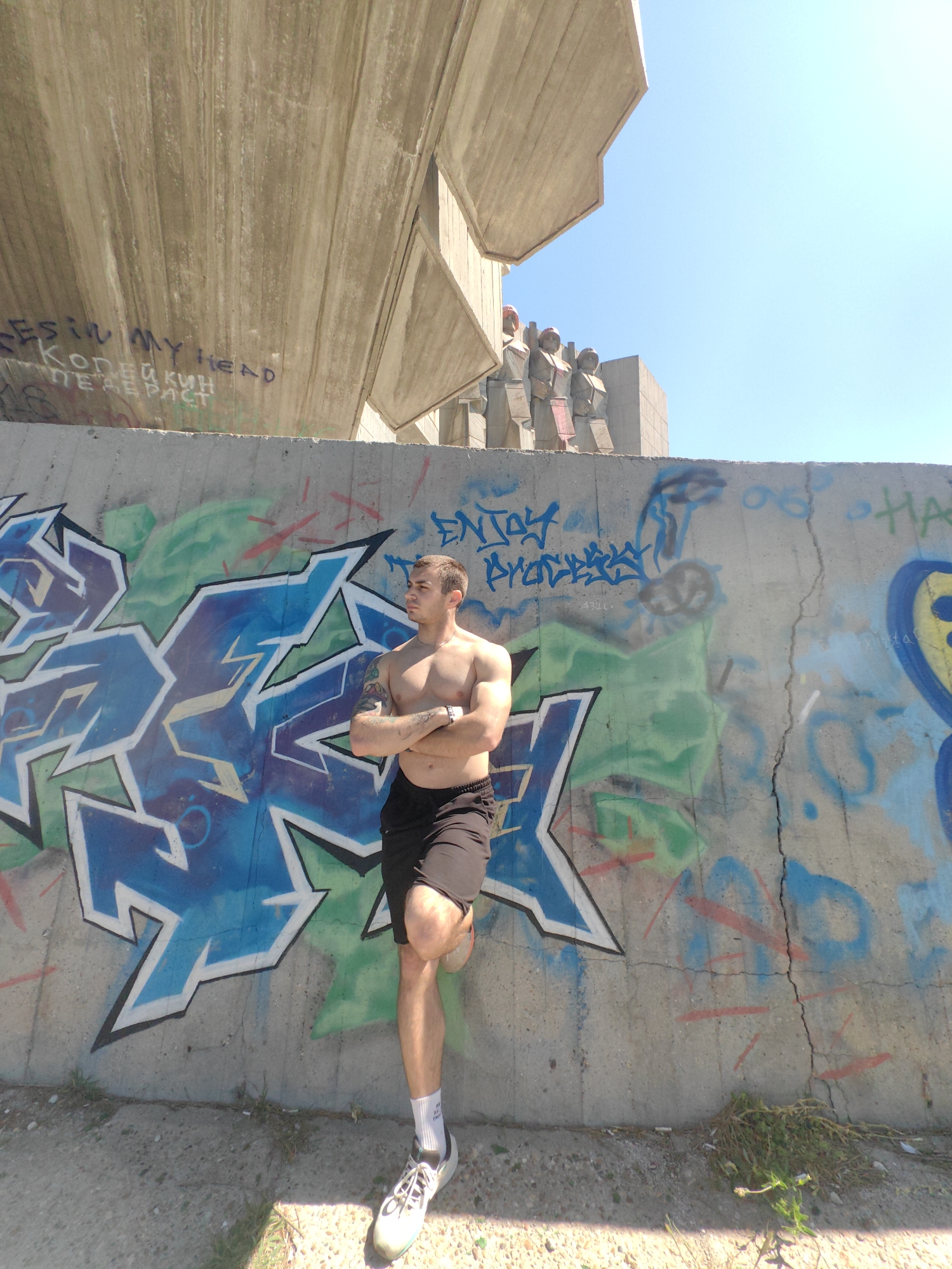 A waist-up shot depicts a muscular man, shirtless and in black shorts, leaning against a concrete wall covered in colorful graffiti. His arms are crossed, and he wears white sneakers and socks. Above the wall, a large, modernist concrete structure looms under a bright blue sky. The location is Unknown, Unknown.