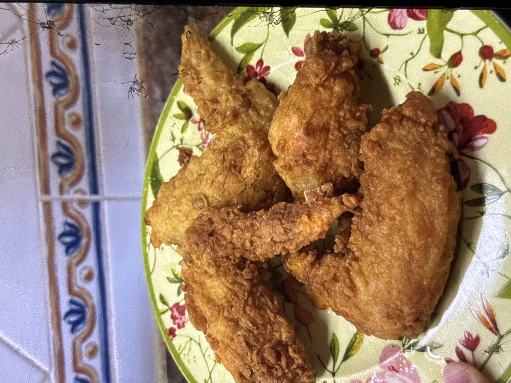 A plate of golden-brown fried chicken wings is presented on a decorative plate with a floral pattern. The plate sits on a tiled surface, possibly a kitchen counter, with a border of blue and brown decorative tiles visible in the background. The lighting suggests an indoor setting, likely a home. The image is a close-up shot, focusing on the food. The location in Orihuela, Spain, is not ascertainable from the visual information provided.