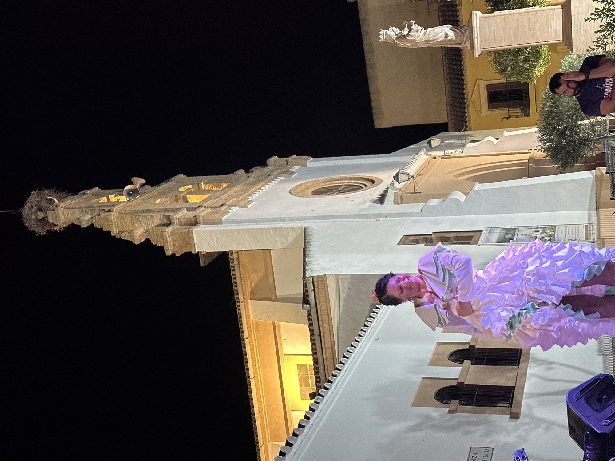 In San José Del Valle, Spain, a woman in a white and pink ruffled flamenco dress performs on a stage at night. She stands before a white building with a terracotta tiled roof and arched windows. Behind her, a tall bell tower with a stork's nest at its apex rises against the dark sky, illuminated by warm interior lights. To the right, a statue of a robed figure stands on a pedestal, and a man in a dark t-shirt is seated nearby. The scene is lit by stage lighting that casts pink and purple hues on the performer and her dress, and a warm yellow light illuminates the building's facade.