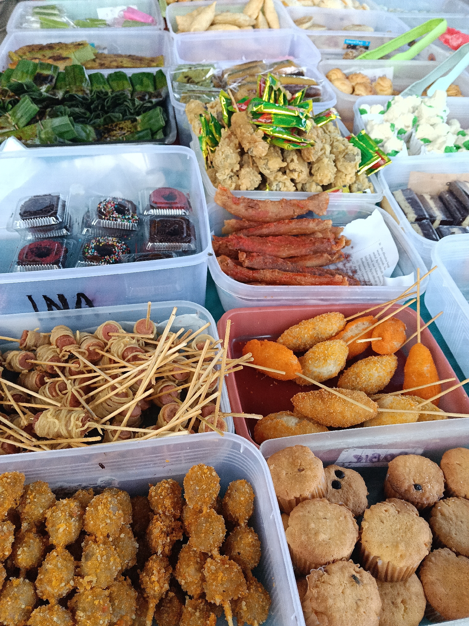 The image shows a food stall, possibly in Lhokseumawe, Indonesia, displaying a variety of local snacks and sweets. The stall consists of clear plastic containers filled with different types of food items.
Visible food includes: green-wrapped snacks (possibly steamed or grilled), donuts, fried sausage-like sticks, fried chicken pieces in colorful wrappers, white cream pastries, chocolate-layered bars, sausage pieces wrapped in noodles on sticks, breaded and fried ball-shaped foods on sticks, and muffins. Some items are on wooden skewers.
The scene appears to be a street-side vending setup. The background is out of focus.