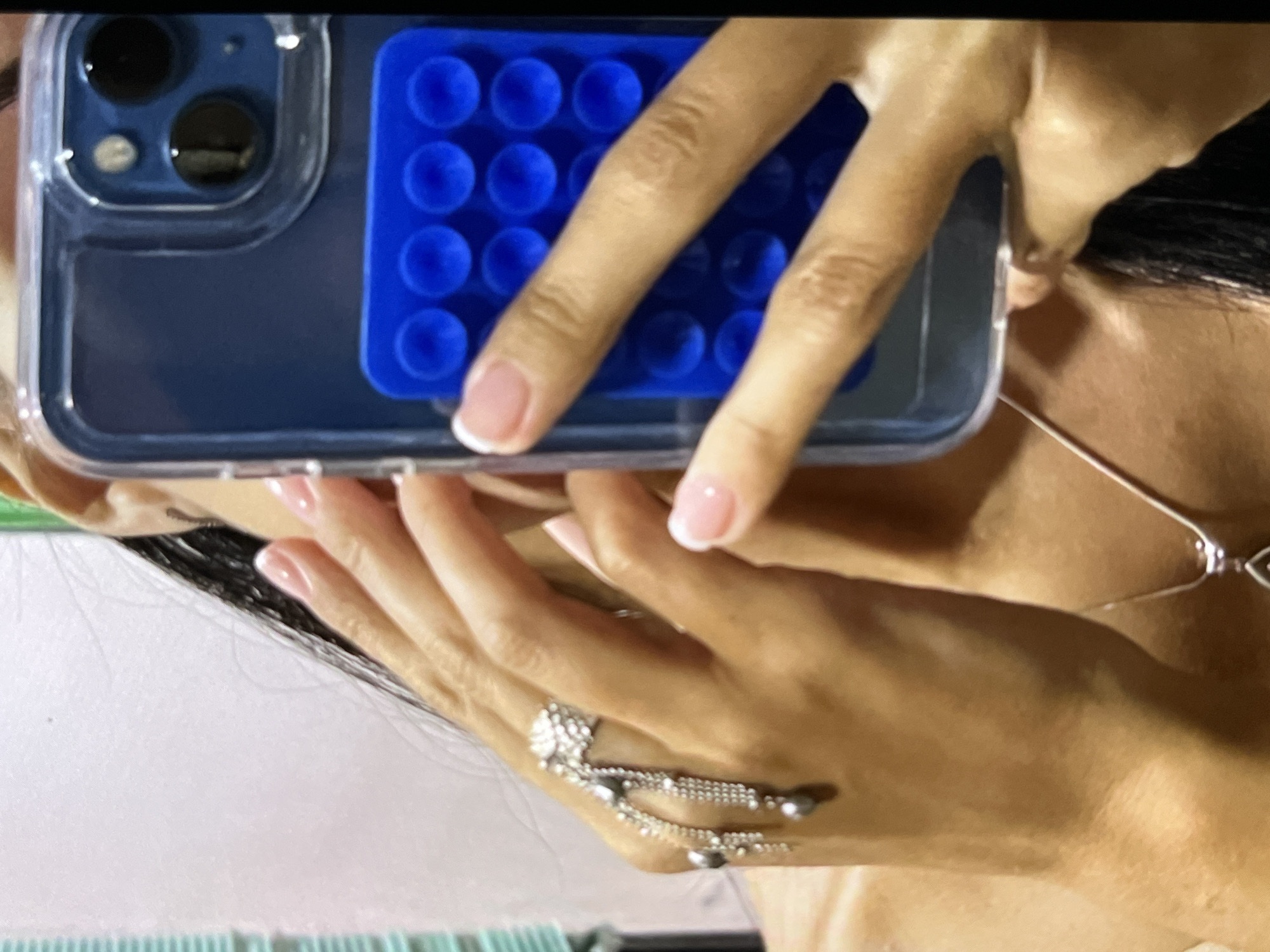 A close-up, angled indoor shot features a person holding a smartphone with a clear case. A blue, rectangular object with multiple circular indentations is adhered to the back of the phone case. The person’s hands are visible, displaying manicured nails with a French tip style. A ring with dangling strands adorns one of the fingers. The background includes portions of the person's face and upper torso. A thin silver necklace is visible. The environment lacks discernible details to determine a specific location within an unknown city in Indonesia.