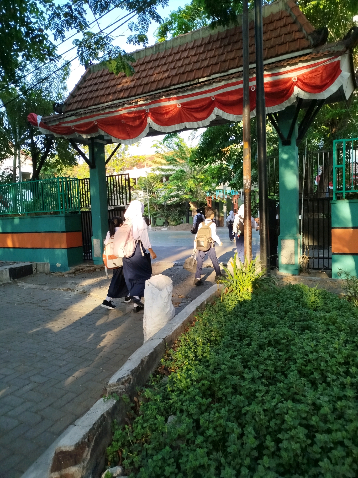 The image depicts the entrance to a school in Surabaya, Indonesia, on a sunny day. The architecture features a tiled roof with red and white decorative banners hanging from the eaves. Two students in uniform are walking away from the camera on a paved walkway. One student, wearing a white hijab and backpack, is walking on the right side of the walkway. Another student, also wearing a hijab, is walking to the left of the first student. A third student, wearing a backpack, is walking ahead of them on the left side of the walkway. The school grounds appear to be surrounded by green fencing and lush vegetation. The overall atmosphere is peaceful and serene, with the warm sunlight casting long shadows on the ground.