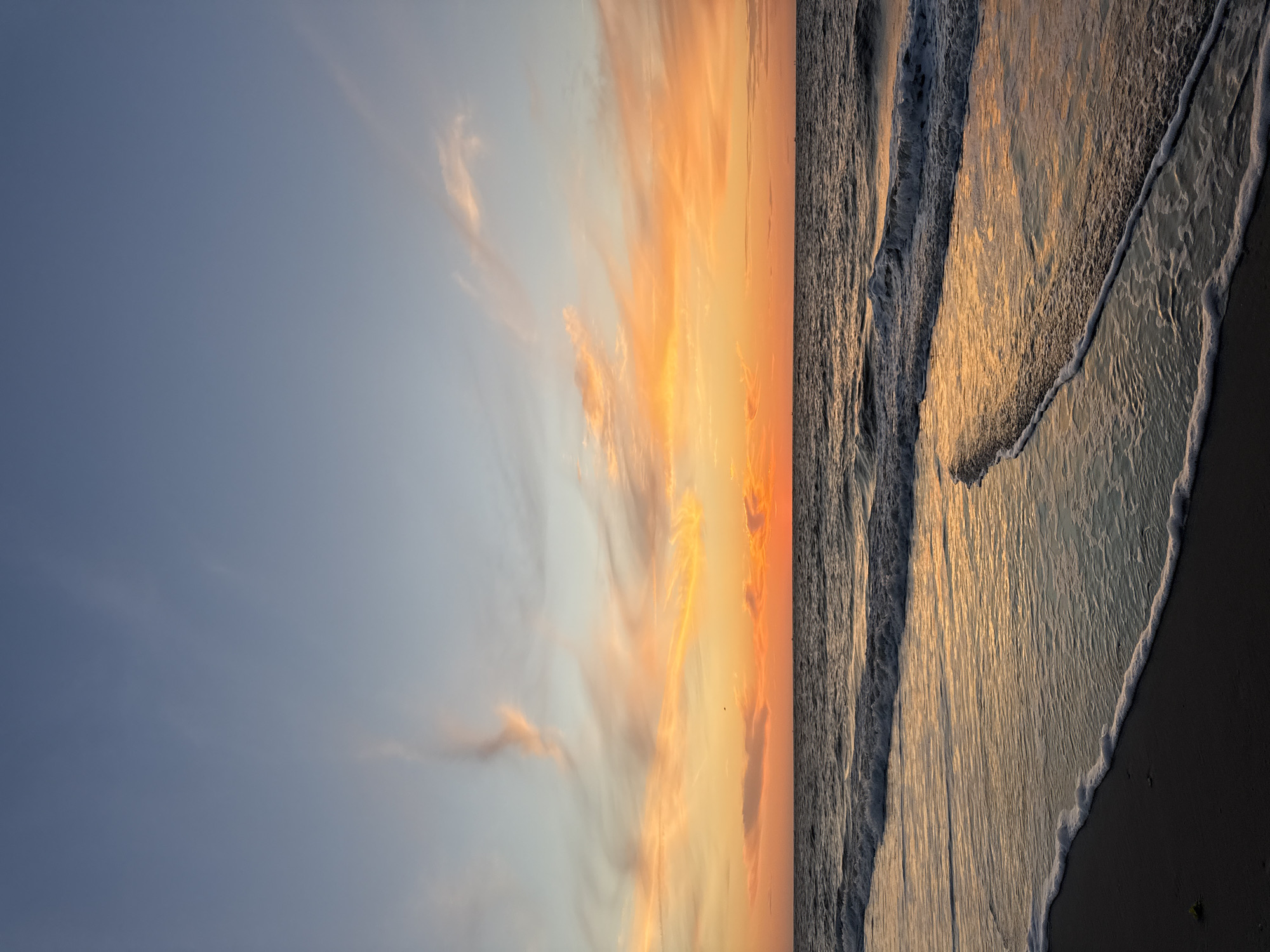 This is a vertical, eye-level, full shot of a sunset at the beach on the island of Langeoog, Germany. The ocean is in the foreground, with waves rolling in towards the shore. The sand is dark and wet, with a line of white foam from the waves. The sky is a gradient of colors, from blue at the top to orange and pink at the horizon, with wispy clouds. The sun is visible as a bright orange orb on the horizon. The overall mood of the image is peaceful and serene.