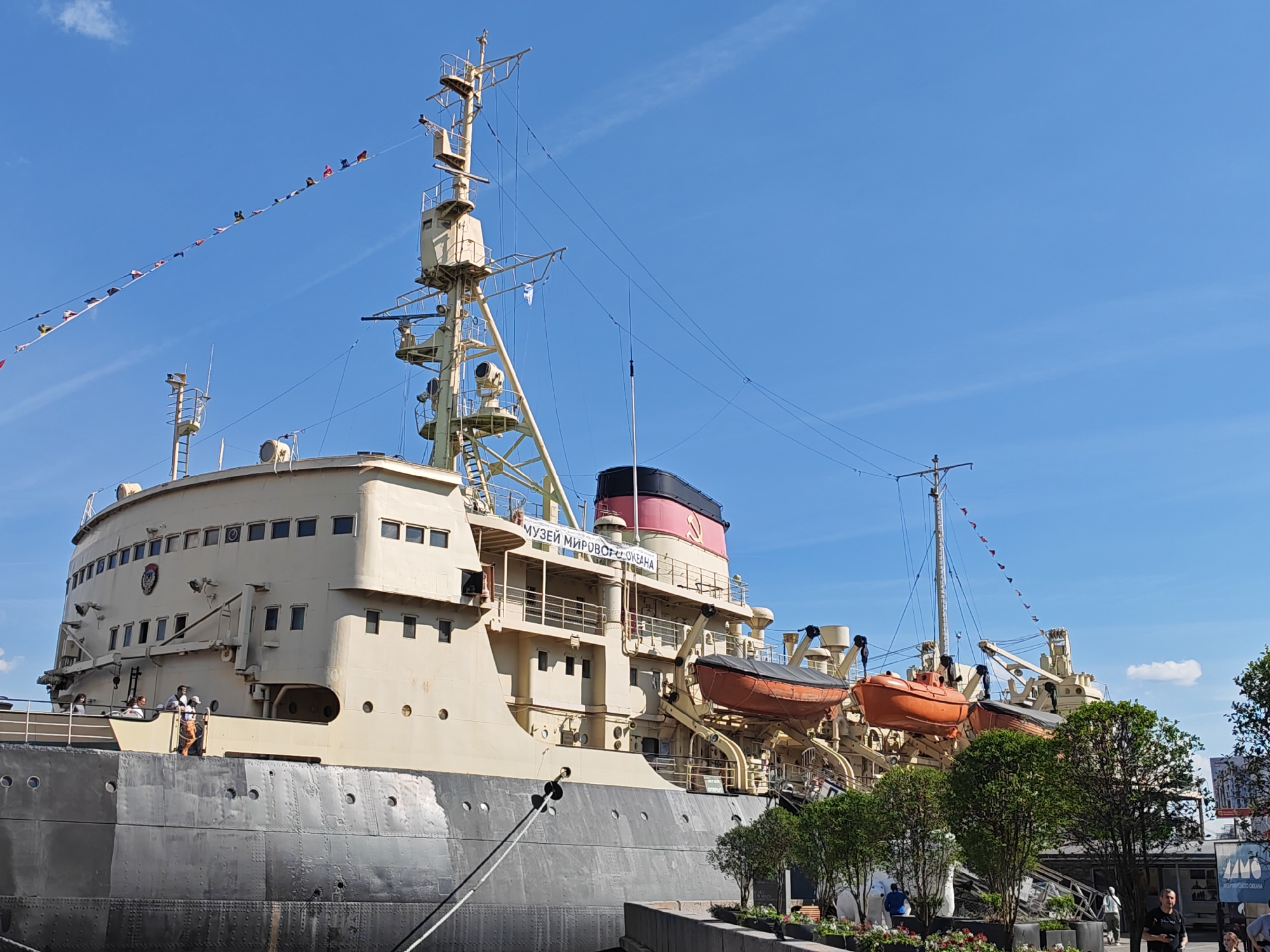 This daytime image captures a large, cream-colored icebreaker ship, identified as a museum, docked under a clear blue sky in what appears to be a port or riverside location in Saint Petersburg, Russia.
The ship, likely the "Lenin" or "Krasin," features a prominent dark grey/black hull with visible rivets and a lighter, beige-colored superstructure. A tall mast with various antennae and radar equipment dominates the upper part of the ship. Decorative nautical flags (bunting) are strung along a line extending from the mast towards the left. Two orange lifeboats are conspicuously mounted on davits on the starboard side of the ship.
Several people are visible: a small group stands on an open deck area of the ship's bow, and others are seen on the paved walkway in the foreground, near lush green trees. Some individuals appear to be tourists or visitors, casually observing the ship.
Notable details include the bright sunlight, indicating a clear, pleasant day. On the side of the superstructure, text in Cyrillic characters reads "МУЗЕЙ МИРОВОГО ОКЕАНА," which translates to "Museum of the World Ocean." The ship's red-topped smokestack bears a faded red star emblem, which, upon closer inspection, appears to include a hammer and sickle, signifying its Soviet-era origin. In the foreground, potted plants and trees line the paved area, with a sign partially visible on the right, also bearing Cyrillic text, possibly related to the museum.