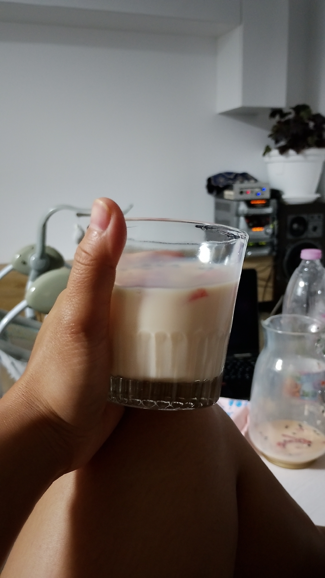 A close-up, eye-level shot shows a person's right hand holding a clear glass filled with a creamy, off-white liquid, possibly milk or a cocktail, with small pinkish-red pieces visible within. The glass has vertical ribbed detailing near its base. The background is blurred, revealing a domestic setting with a white wall, a shelf unit with electronic equipment, speakers, and a potted plant. To the right of the glass, a clear glass pitcher containing a similar liquid and possibly some sediment is partially visible. The person's thumb is extended upwards, while their index and middle fingers are curled around the glass. The image appears to be taken indoors, likely in a living space or bedroom. There is no identifiable location in Smolyan, Bulgaria.