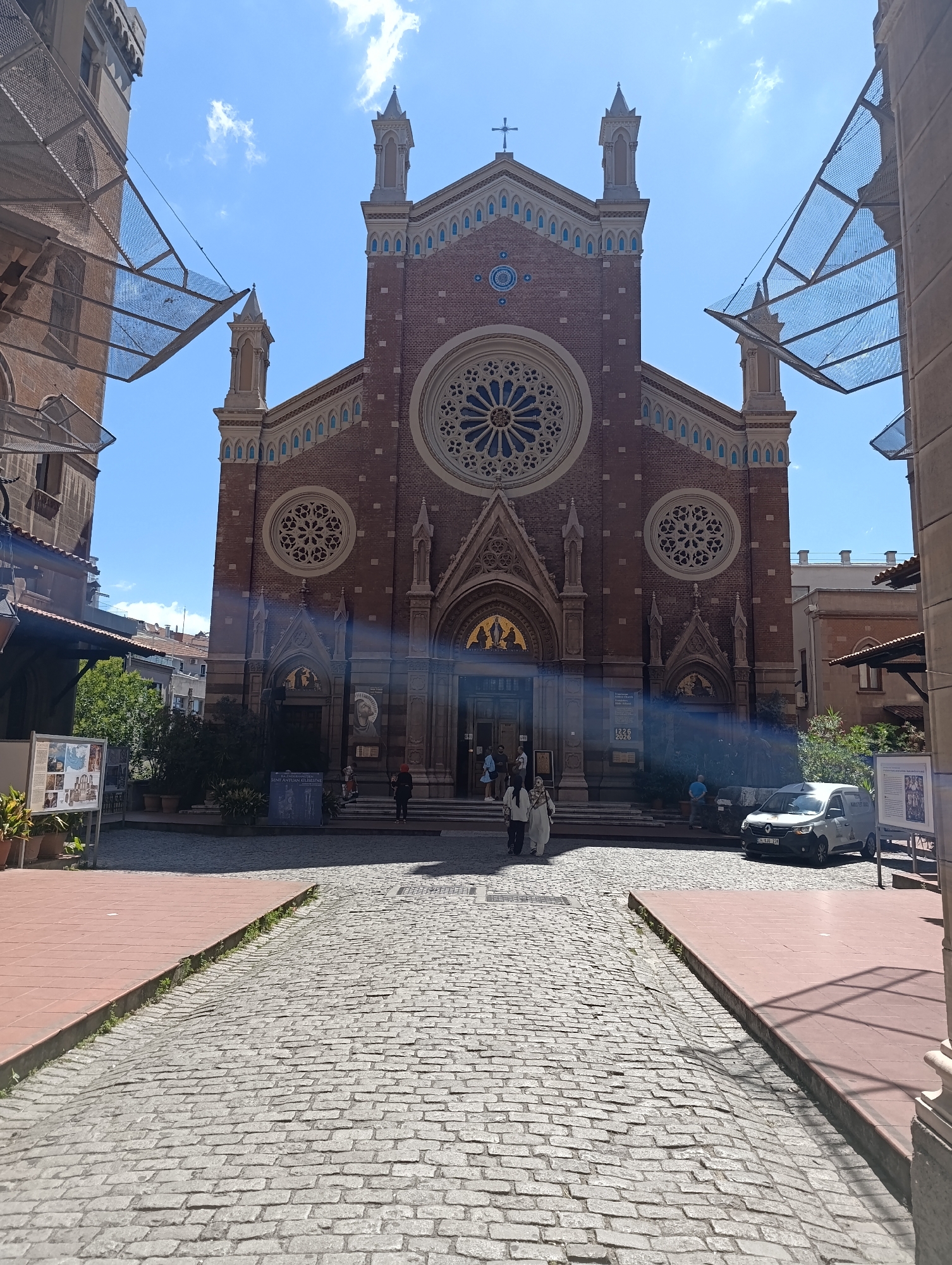 This image depicts the exterior of St. Anthony of Padua Church in the Beyoğlu district of Istanbul, Turkey, on a clear, sunny day. The church, constructed of red brick with Gothic Revival architectural elements, features a large rose window above its entrance and two smaller circular windows on either side. The main entrance is a grand arched doorway with intricate detailing.
In front of the church, a cobblestone pathway leads towards the entrance. The pathway is bordered on both sides by brick-paved areas and some planters with greenery. A few individuals are visible on the steps of the church and walking along the pathway. To the right, a white van is parked.
The scene is framed by buildings on either side, one appearing to be part of the church complex and another a more modern structure. Overhead, there are some architectural elements that appear to be decorative canopies or sunshades made of mesh. The sky is blue with a few scattered clouds. Information panels are placed near the entrance, suggesting a tourist or informational aspect to the location.