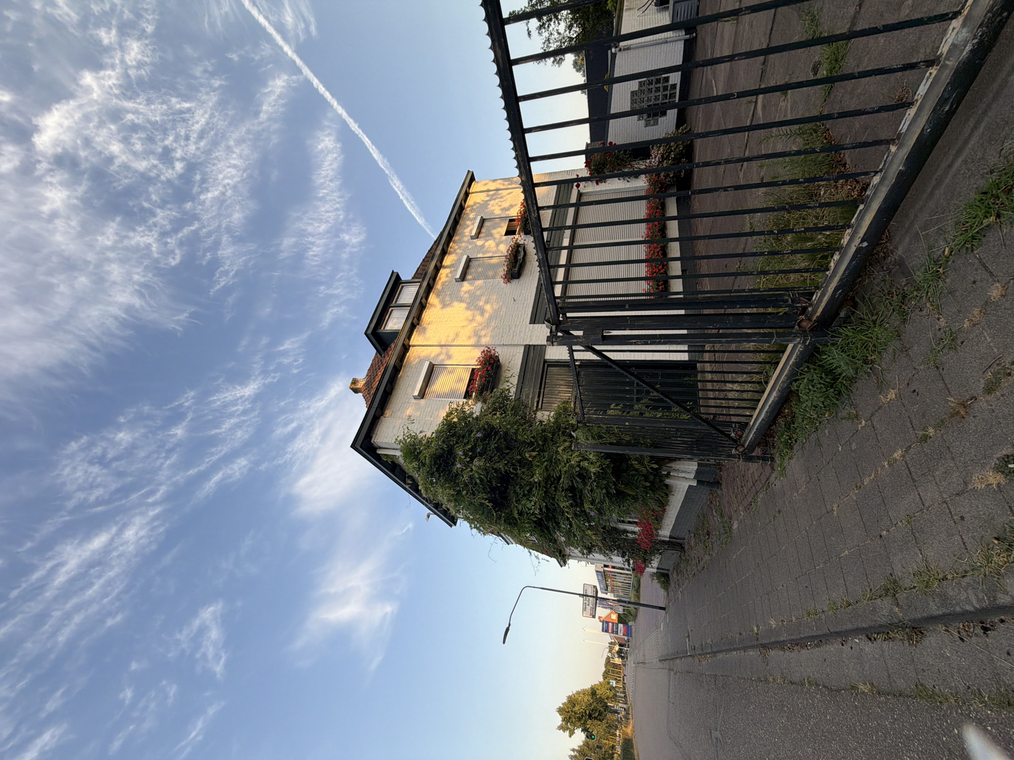 The image shows a white brick building with a dark green trim and a tiled roof, overgrown with ivy on its left side. The building has several windows, some covered by white rolling shutters. Flower boxes with red flowers are visible below some of the windows. A black metal fence with vertical bars encloses the front of the property. To the left of the building is a paved sidewalk and a road. A lamppost stands on the sidewalk. In the background, there are some trees and signs, suggesting an urban or suburban setting. The sky is blue with wispy white clouds and a contrail from an aircraft. This image was taken on the Kanaaldijk N.W. in Helmond, Netherlands, near the intersection with the West-Koninginneweg, a location characterized by its proximity to both residential and commercial areas along the canal. There are no people visible in the scene. The overall impression is one of a property with a rustic, natural element (the ivy) integrated into a more built environment.