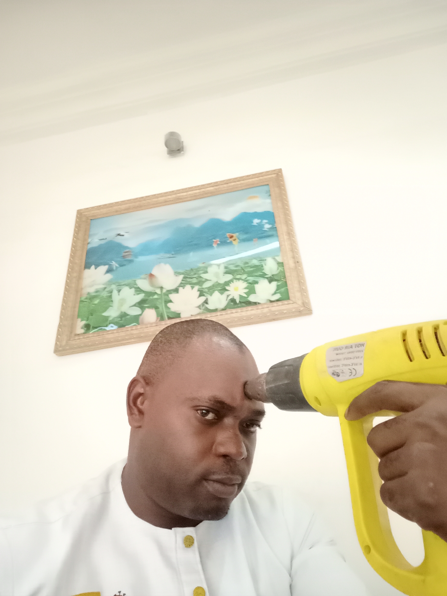 The image features a dark-skinned man, captured indoors in what appears to be Gwarinpa, Nigeria. He is seen in a close-up, posing with a bright yellow heat gun pointed directly at his forehead. The man, with short dark hair, wears a white garment featuring golden buttons and embroidery, and looks intensely at the viewer with a serious or resolute expression. His right hand is visible, gripping the handle of the heat gun.
On the side of the yellow tool, text such as "HOT AIR GUN" is visible, along with a "CE" certification mark and other smaller, indistinct lettering. The background consists of plain white walls, suggesting a simple interior setting. A framed painting hangs on the wall behind and to the left of the man. This artwork depicts a serene landscape with a prominent foreground of large white water lilies or lotus flowers, a body of calm blue water, distant mountains, and a clear blue sky with a few birds or flying insects. A small, contemporary wall-mounted light fixture is also visible on the white wall above the painting. The overall lighting is bright and even, indicating daytime. The pose with the tool suggests a symbolic or dramatic gesture rather than a literal action.
