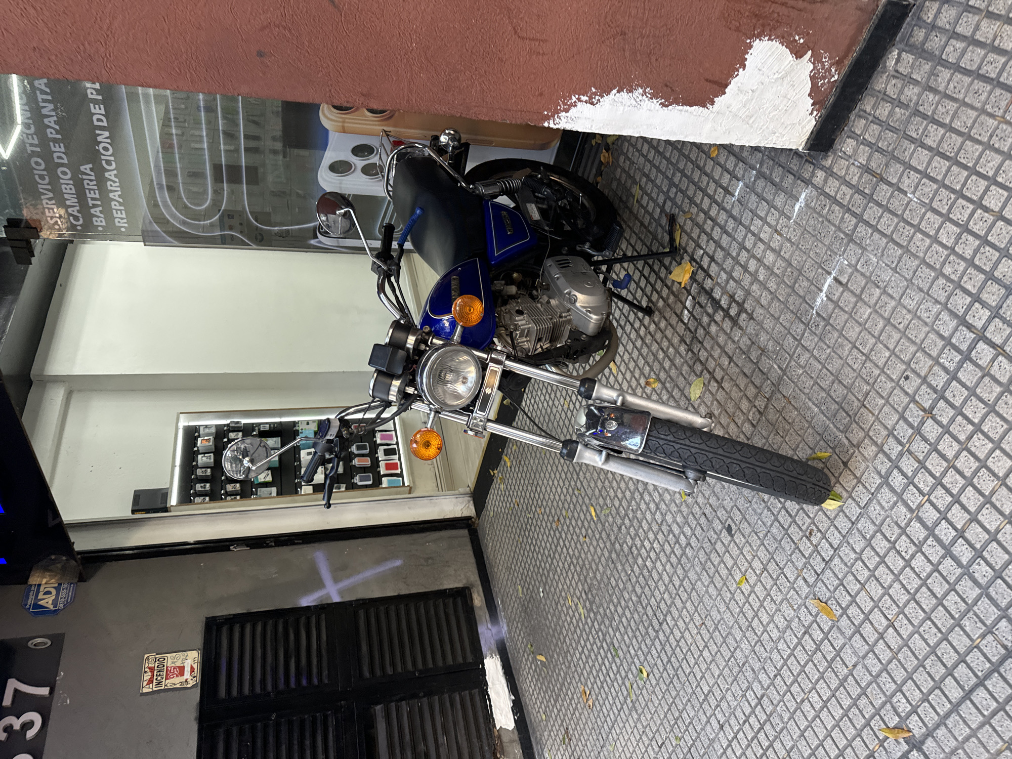 The image shows a blue and silver motorcycle parked on a grey tiled sidewalk in front of a building. The building has a red wall at the top and a grey wall at the bottom. The building houses a business offering technical services for cellphones and batteries. The motorcycle is parked close to the wall, with its front tire facing the camera. Some leaves are scattered on the sidewalk. The street number "37" is visible on the building. There is a sign "Incendio" on the wall, indicating fire equipment or instructions. "0810-555-10" and "ADT" stickers are also affixed to the wall. Based on the Spanish signs, the location is likely in Buenos Aires, Argentina.