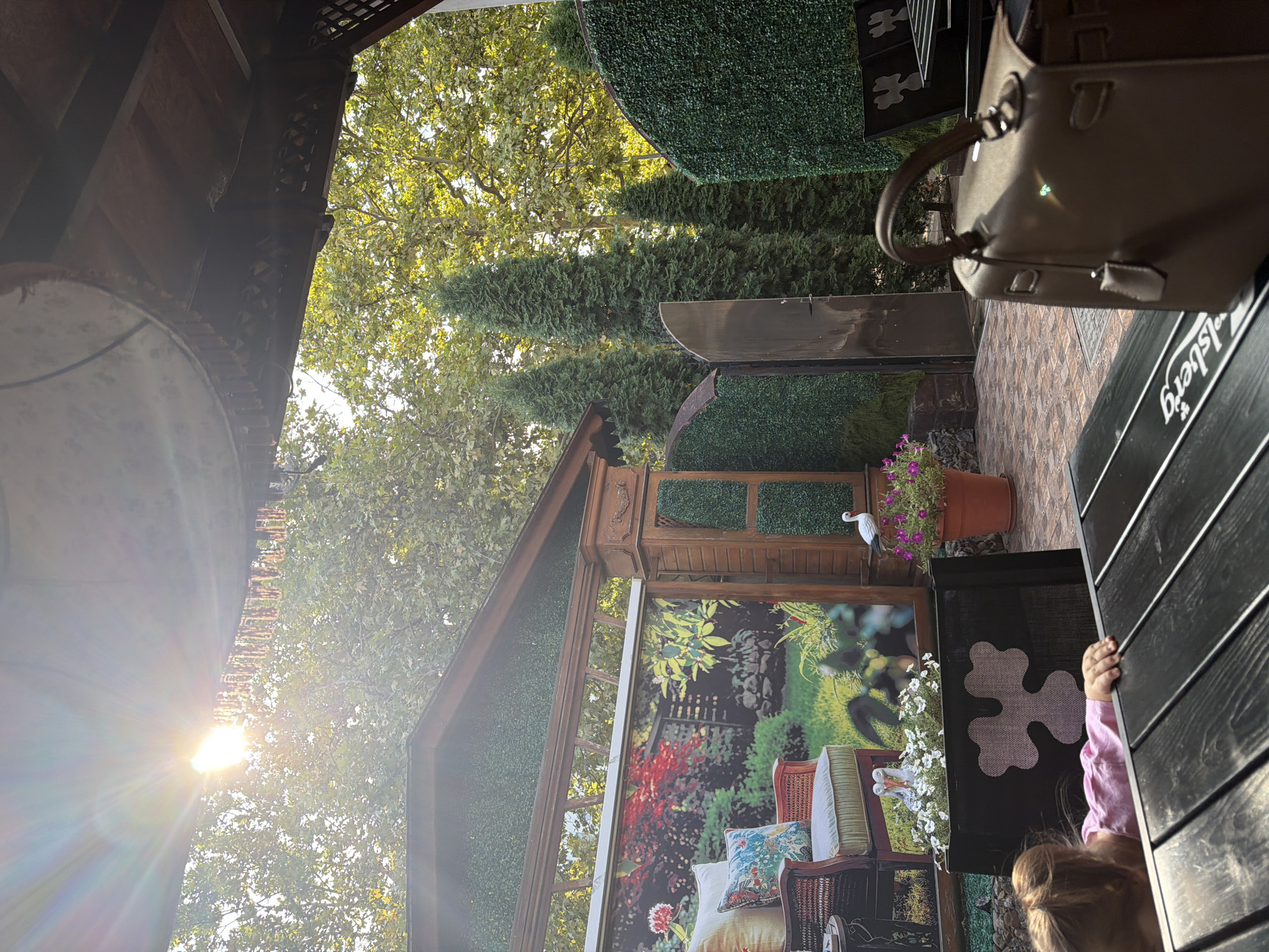 The image depicts an outdoor seating area, likely a cafe or restaurant, with a rustic and naturalistic aesthetic. Sunlight streams in from the upper left, creating lens flare and highlighting the green foliage of trees in the background. In the foreground, a dark wooden table is visible, with a brown leather handbag placed on it. To the left, a young child, wearing a pink shirt, is reaching towards the table. Behind the table, there is a decorative black screen with a white clover-like pattern.
Further into the scene, a backdrop featuring a printed garden scene is displayed on a wooden structure. This backdrop shows a bench with cushions, potted plants, and a fence. To the right of the backdrop is a wooden pillar and an area covered in artificial green turf. Tall cypress trees and more artificial turf walls create a sense of enclosure. A metal gate is also visible within the artificial turf wall. On the right side of the image, more outdoor seating with chairs and tables is apparent, also adorned with the clover-like pattern on the backrests.
The environment suggests a warm, possibly late afternoon setting, with dappled sunlight filtering through the leaves. The presence of a printed garden backdrop and artificial turf points towards a cultivated outdoor space designed for patrons. While the exact location within Soroca, Moldova, is not explicitly stated, the scene suggests an establishment that aims to create a relaxing and visually appealing outdoor ambiance.
