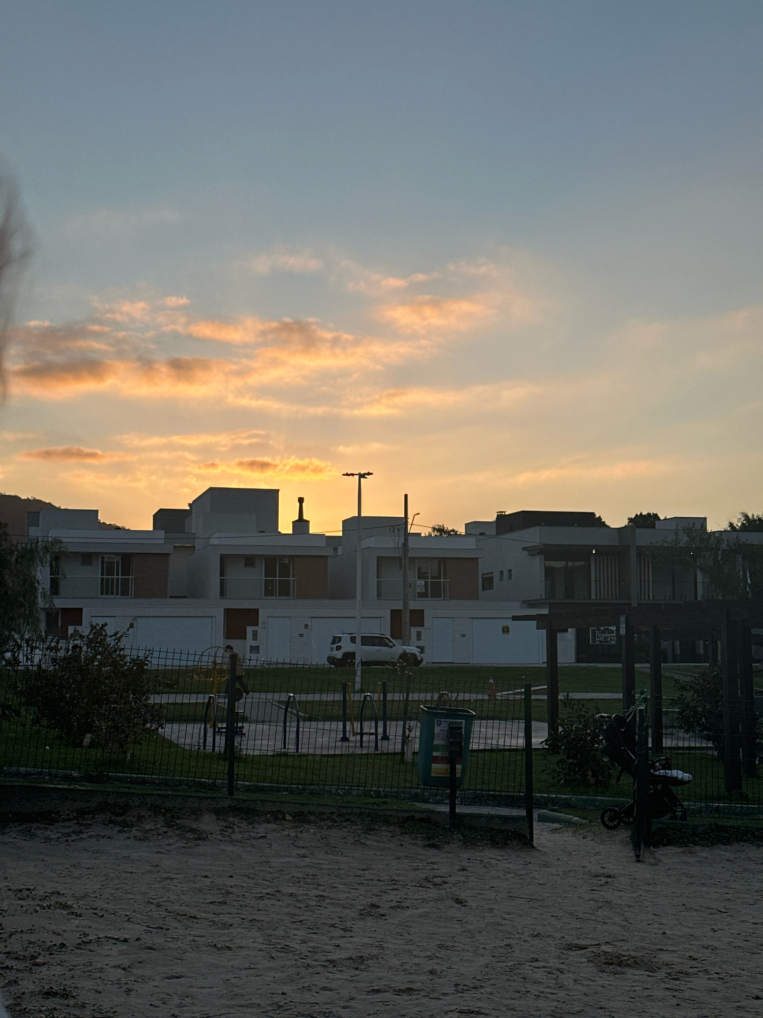 A dusk sky with orange and pink clouds is visible above a row of modern, multi-story houses. A playground with metal equipment and a trash can is in the foreground, separated from the houses by a fence. A white SUV is parked in front of one of the houses. The ground in the immediate foreground is sandy.