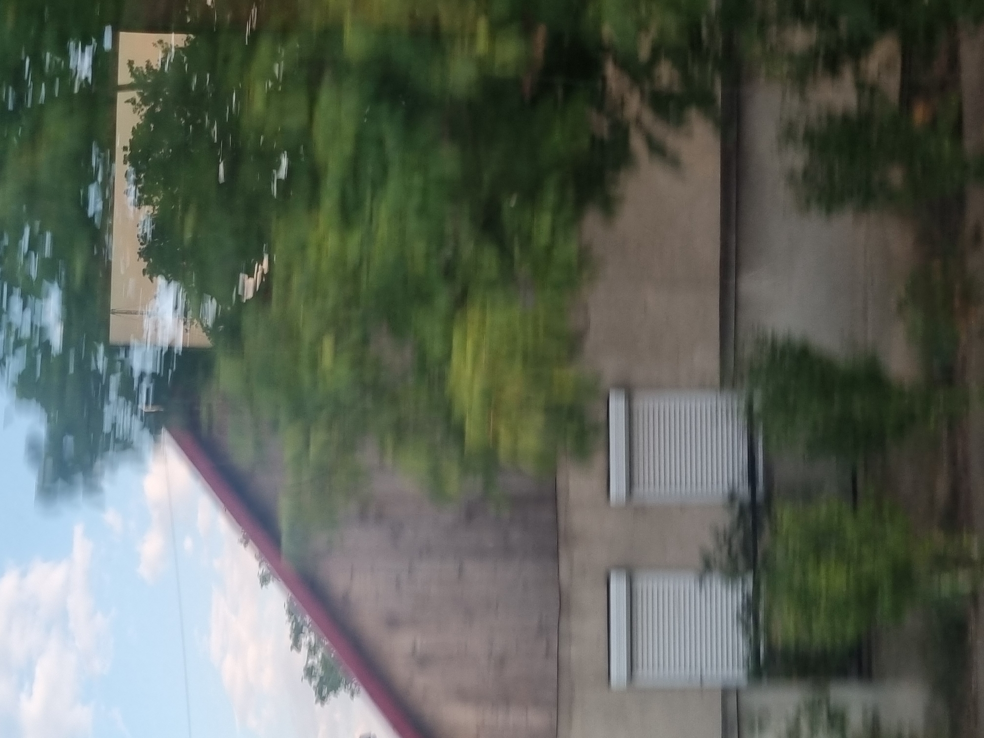 The image depicts a blurred, motion-captured view of a building facade, likely taken from a moving vehicle. The building appears to be two stories high. The upper portion is clad in wooden siding with a red-tiled roof. Below this, the facade is a neutral color, possibly concrete or stucco. Two windows, each covered with white roller shutters, are visible on the lower level. Green foliage, suggesting trees or shrubs, is present in the foreground and also partially obscures the building. The sky, with scattered clouds, is visible in the upper left portion of the image. The overall impression is one of a rural or semi-rural setting, possibly on the outskirts of a town or village. The location is identified as Gepiu, Romania.