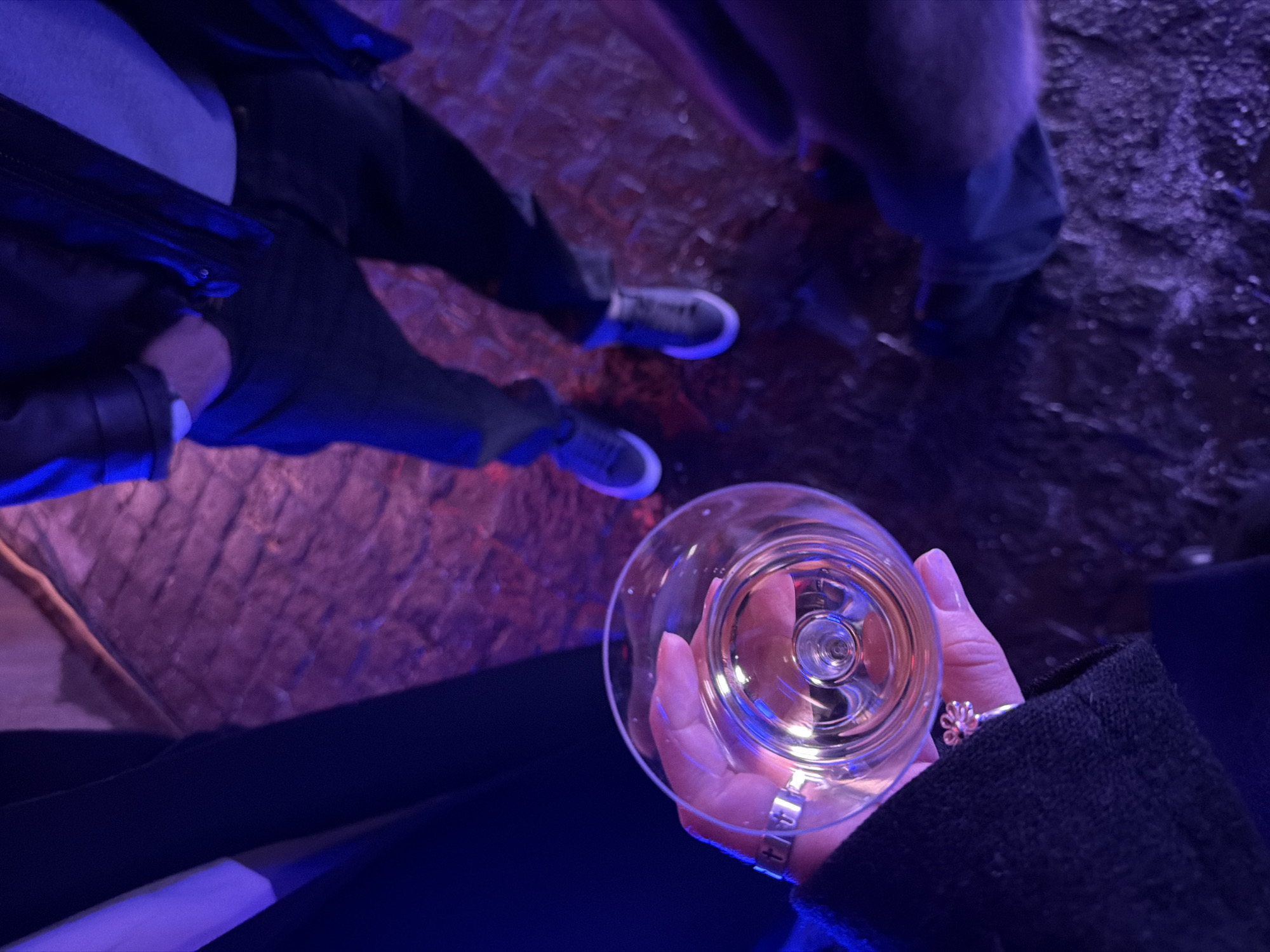 A low-angle shot depicts a woman's hand holding a clear wine glass filled with a light-colored liquid. The woman wears a ring and a bracelet on the hand holding the glass. The background shows people's legs and feet standing on a surface that appears to be textured, potentially a rough floor or even a wall texture. The overall lighting has a blue and purple hue, which adds to the atmosphere. The specific location is not identifiable from this image without additional information, and there is no evidence to suggest the image was taken in Moscow, Russia.