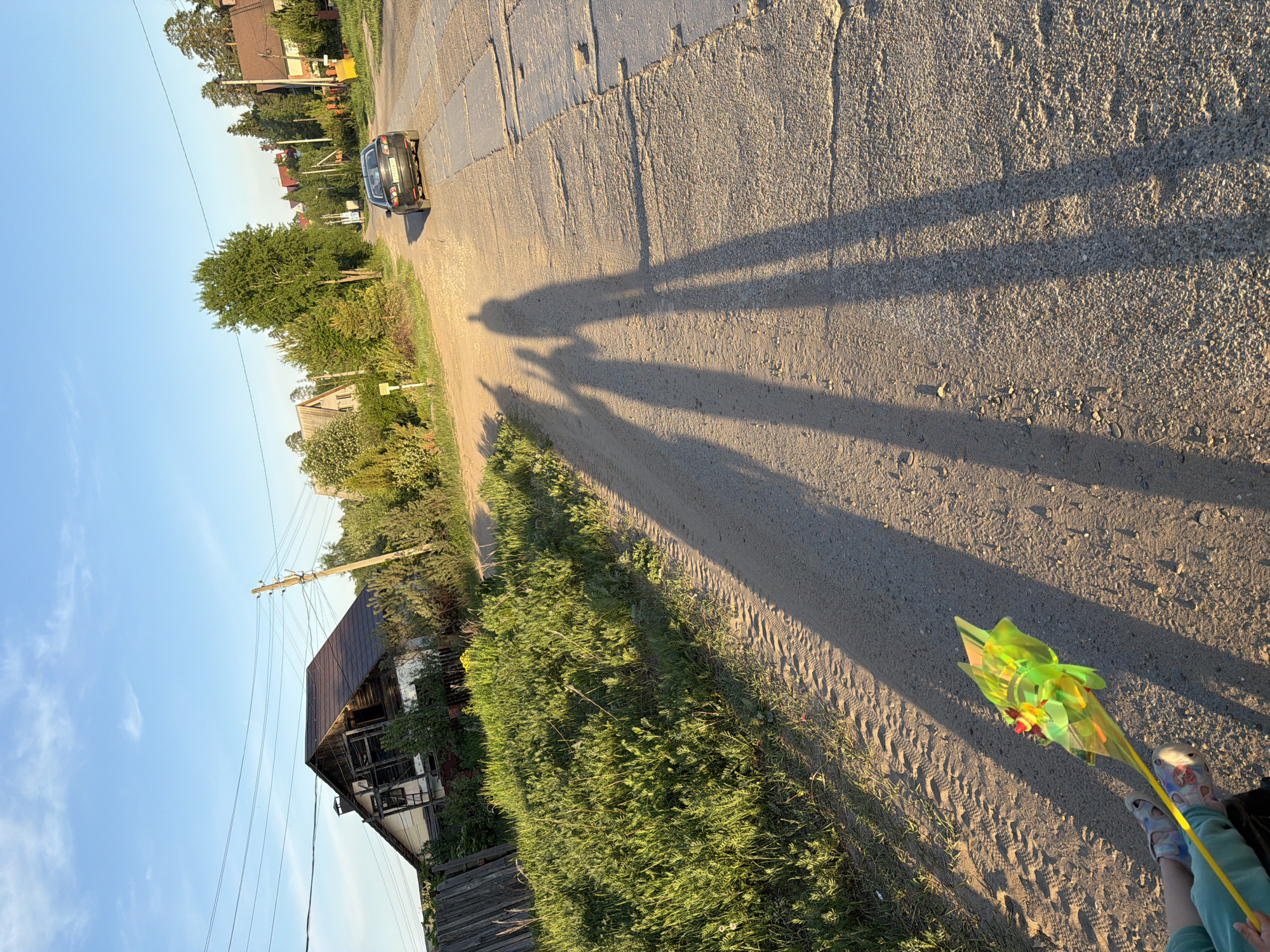 The image shows a street scene with a car visible in the distance on the left, and shadows of people cast onto the road. The road appears to be cracked and uneven. Along the side of the road, there is overgrown vegetation and an old, dilapidated wooden building with a dark roof. Power lines and a utility pole are present near the building. In the foreground, a child's legs are visible, holding a yellow stick with a green and yellow decorative object attached to it. The sky is blue and the lighting suggests it is late afternoon or early evening. The location is likely a rural or suburban area in Chaikovsky, Russia, given the architectural style of the building and the general environment.