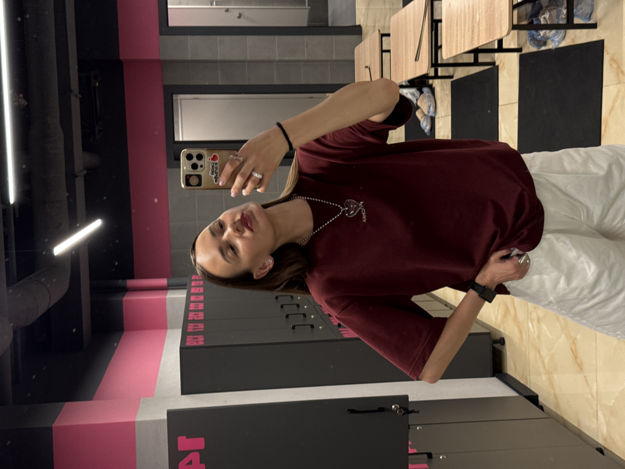 A young woman takes a mirror selfie in what appears to be a locker room, possibly at a gym or sports facility in Sochi, Russia. She is wearing a maroon t-shirt and white shorts. She has on a necklace with a heart-shaped pendant, a watch on her left wrist, and rings on her fingers. Her phone, in a beige case with stickers, is held up to take the photo. The background shows rows of lockers, wooden benches, and a tiled floor. The color scheme of the room includes pink and gray accents on the walls.