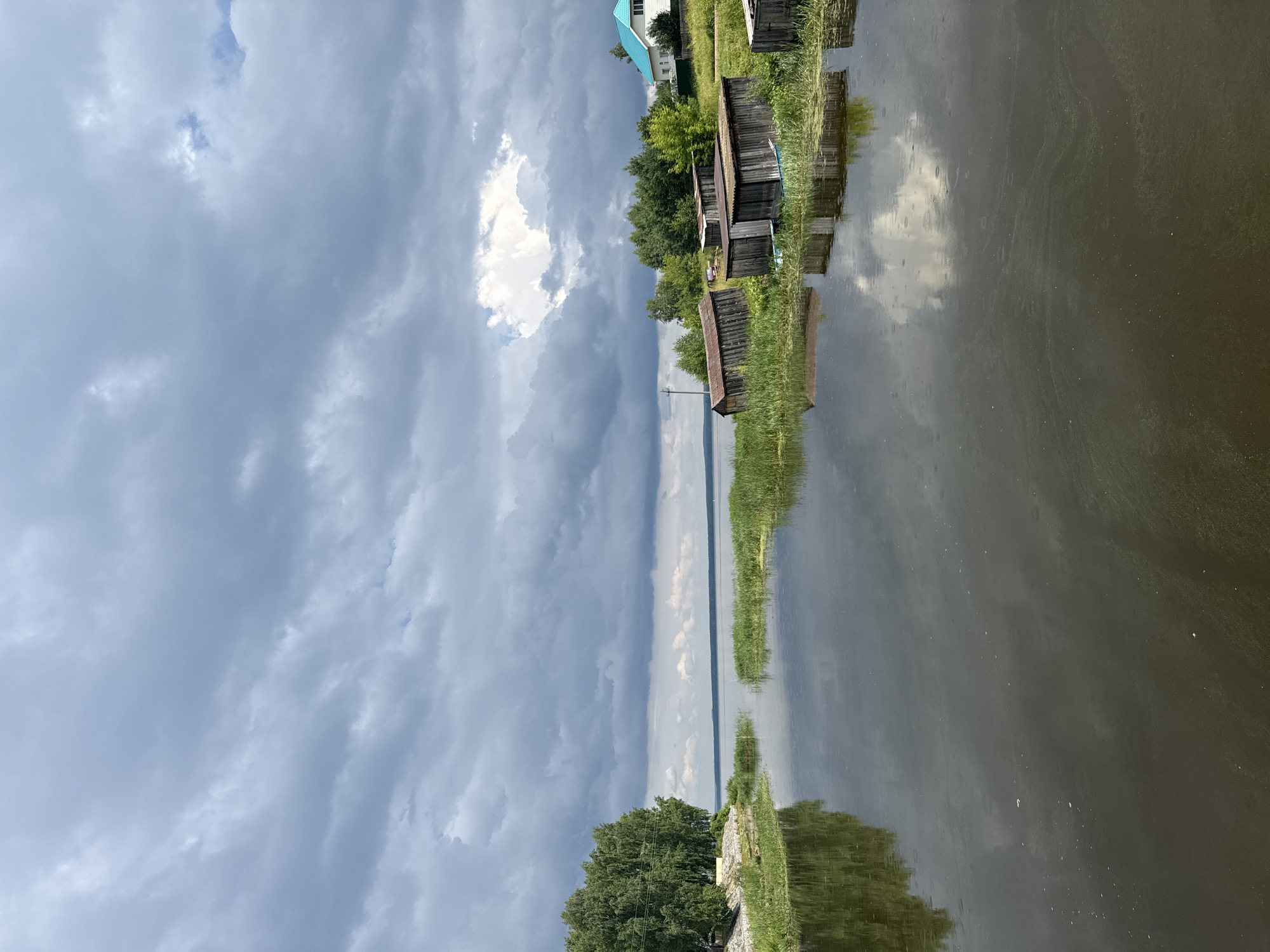 The image depicts a tranquil lake scene in Chastyye, Russia, under a dramatic, cloudy sky. The foreground is dominated by dark, reflective water, with a line of tall green reeds dividing the water's surface. To the left, a cluster of lush green trees stands on a rocky bank, their reflection mirroring in the water. Along the right bank, several weathered wooden structures, appearing to be boathouses or storage sheds, are situated amongst the reeds. In the distance, across the expansive lake, a small settlement is visible, featuring a white house with a bright turquoise roof, nestled amongst more trees. The overall atmosphere is one of serene, natural beauty, tinged with a hint of melancholy due to the overcast sky. There are no people or animals visible in the frame.