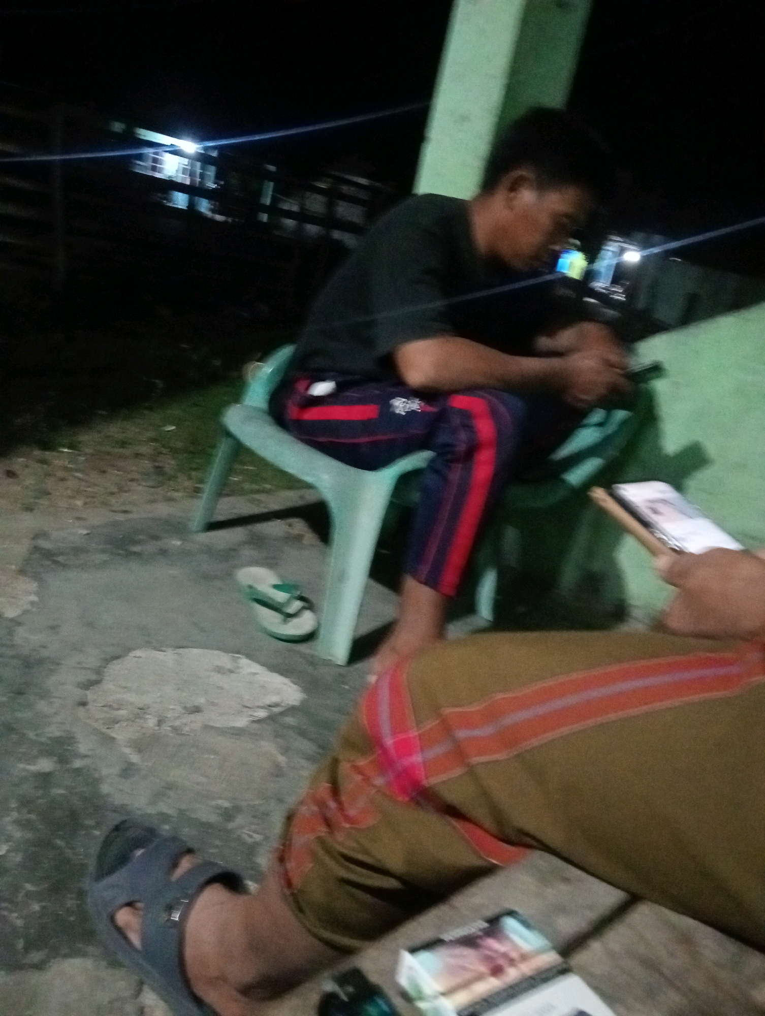 In a nighttime outdoor setting, likely in Tanjung Pandan, Indonesia, two individuals are partially visible. One person, seated on a light blue plastic chair, wears a dark t-shirt and dark pants with red stripes. This individual appears to be holding a dark-colored mobile phone. A single green flip-flop is visible on the ground near the chair.
The other person's legs and feet are in the foreground. They are wearing a patterned sarong or similar garment in earthy tones with horizontal stripes of red and lighter shades. A grey sandal is on their left foot.
In front of this second individual, on a wooden surface, lies a pack of cigarettes. The background is dark, with faint outlines of structures and a bright light source visible in the distance, suggesting artificial lighting from a building. A green column, possibly part of a structure, is on the right side of the image. The ground appears to be a mixture of concrete and dirt with some gravel.