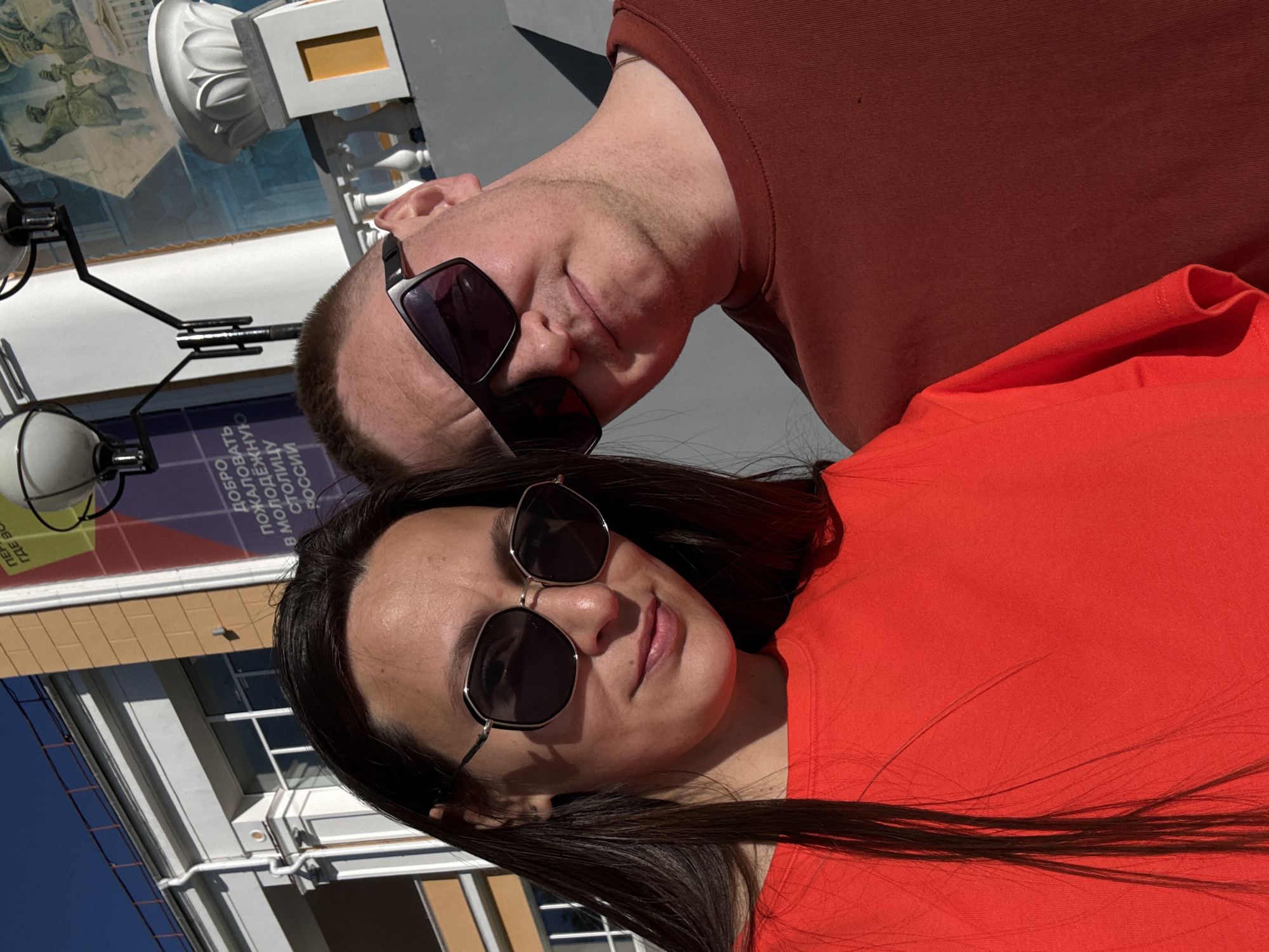 A selfie shows a woman with dark hair and sunglasses and a man with sunglasses. The woman is wearing an orange shirt and the man a maroon shirt. They are in Perm, Russia, near a building with a white balustrade and mural. A streetlight fixture is visible.