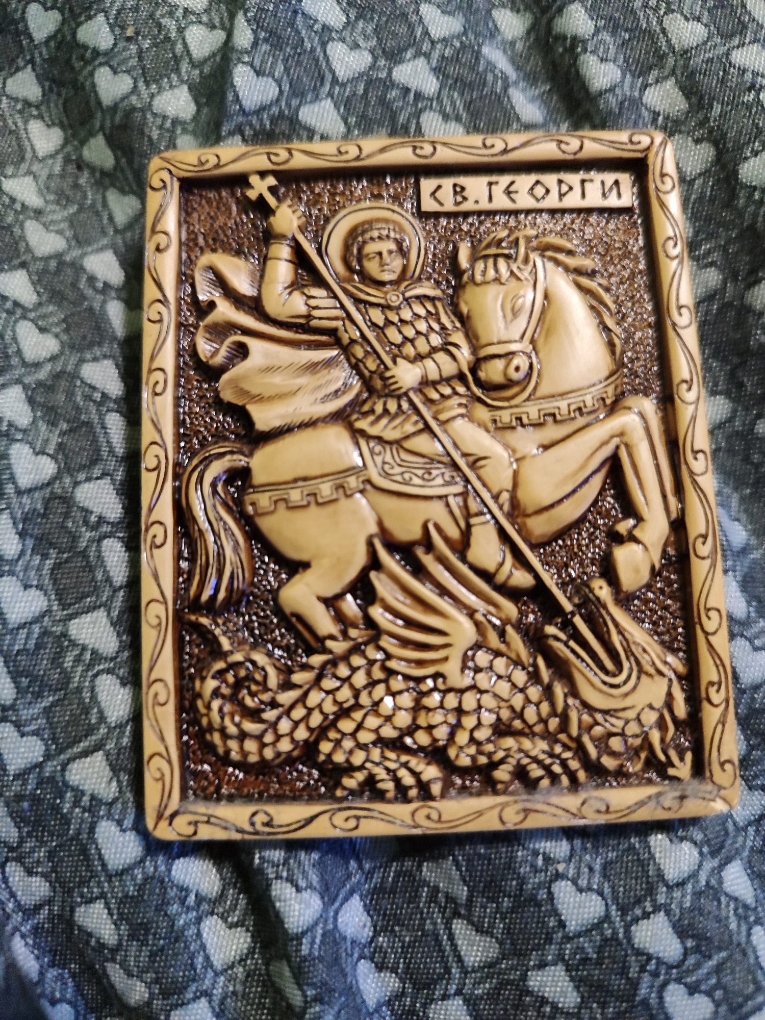 The image shows a rectangular, carved wooden plaque depicting Saint George slaying the dragon. The plaque has a decorative border with swirling patterns. Saint George is portrayed on horseback, holding a spear pointed at a dragon beneath the horse. Above the figures, there is an inscription in Cyrillic, likely identifying the saint. The plaque rests on a patterned fabric with small, repeating heart shapes. This image appears to be a depiction of Saint George from Bulgaria as the inscription above Saint George is written in the Bulgarian Cyrillic alphabet. Burgas, Bulgaria is known for its Bulgarian Orthodox Christian Churches and iconography, so this wooden plaque may be a commemorative icon depicting St. George and The Dragon. There are no visible persons in the image other than the figure of Saint George.