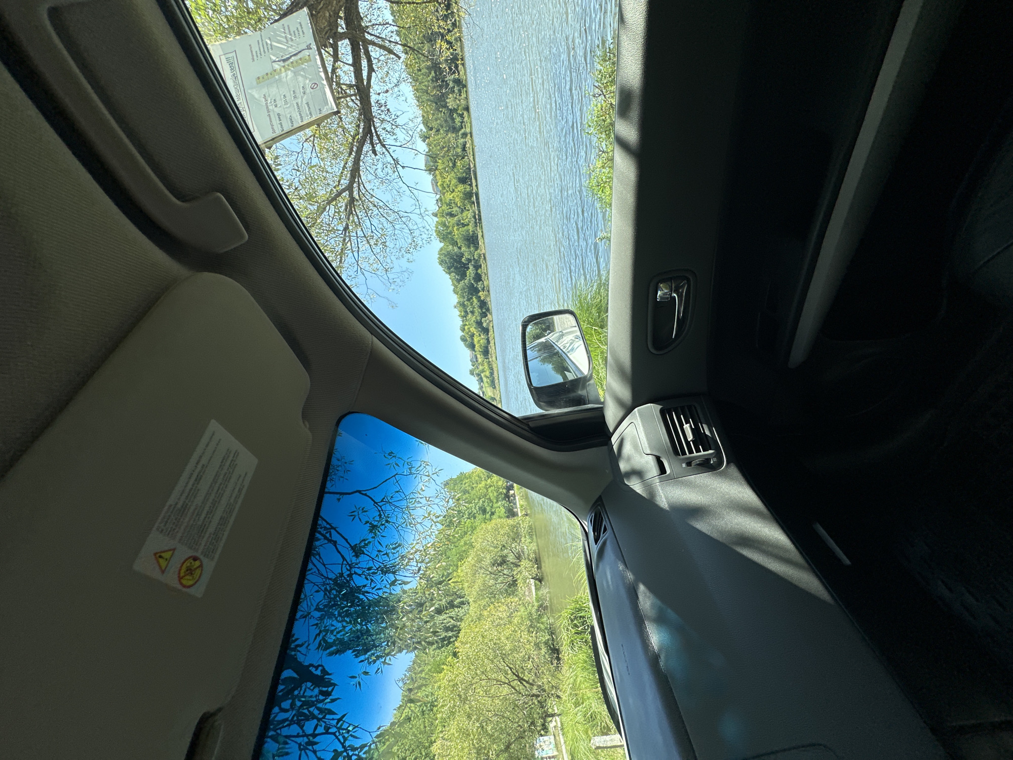 The image is taken from the interior of a vehicle, looking out the driver's side window. The vehicle is parked on a grassy area next to a body of water, likely a river or lake, with lush green trees and vegetation on the opposite bank. The sky is clear and blue, indicating a sunny day. In the foreground, the dashboard and door panel of the car are visible. A sun visor with warning labels is also present on the roof. On the right side of the image, a side mirror reflects the surroundings. In the distance, across the water, a gently sloping hill covered in trees can be seen, with some houses visible towards the top. The overall scene suggests a peaceful, natural setting. The mention of Makiv, Ukraine, implies this natural scene is located within or near that city.