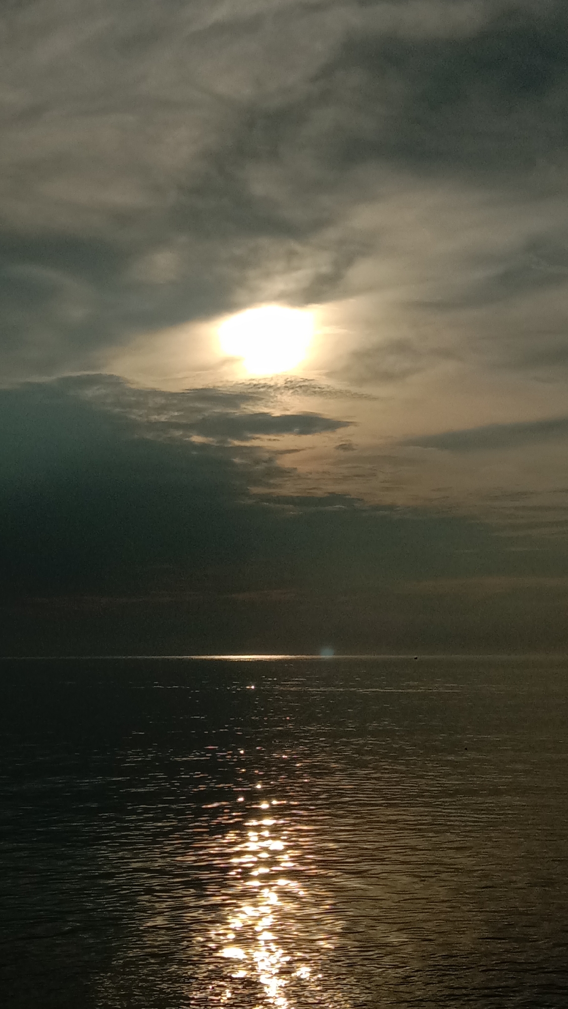 The image shows a view of the sea at sunset or sunrise, likely in Lhokseumawe, Indonesia. The sun, partially obscured by clouds, casts a bright glow on the water. Reflections of the sun create a shimmering effect on the water's surface. The sky is filled with dark, heavy clouds. There is a distinct horizon line separating the sky from the sea. There are no visible people or other objects in the image.