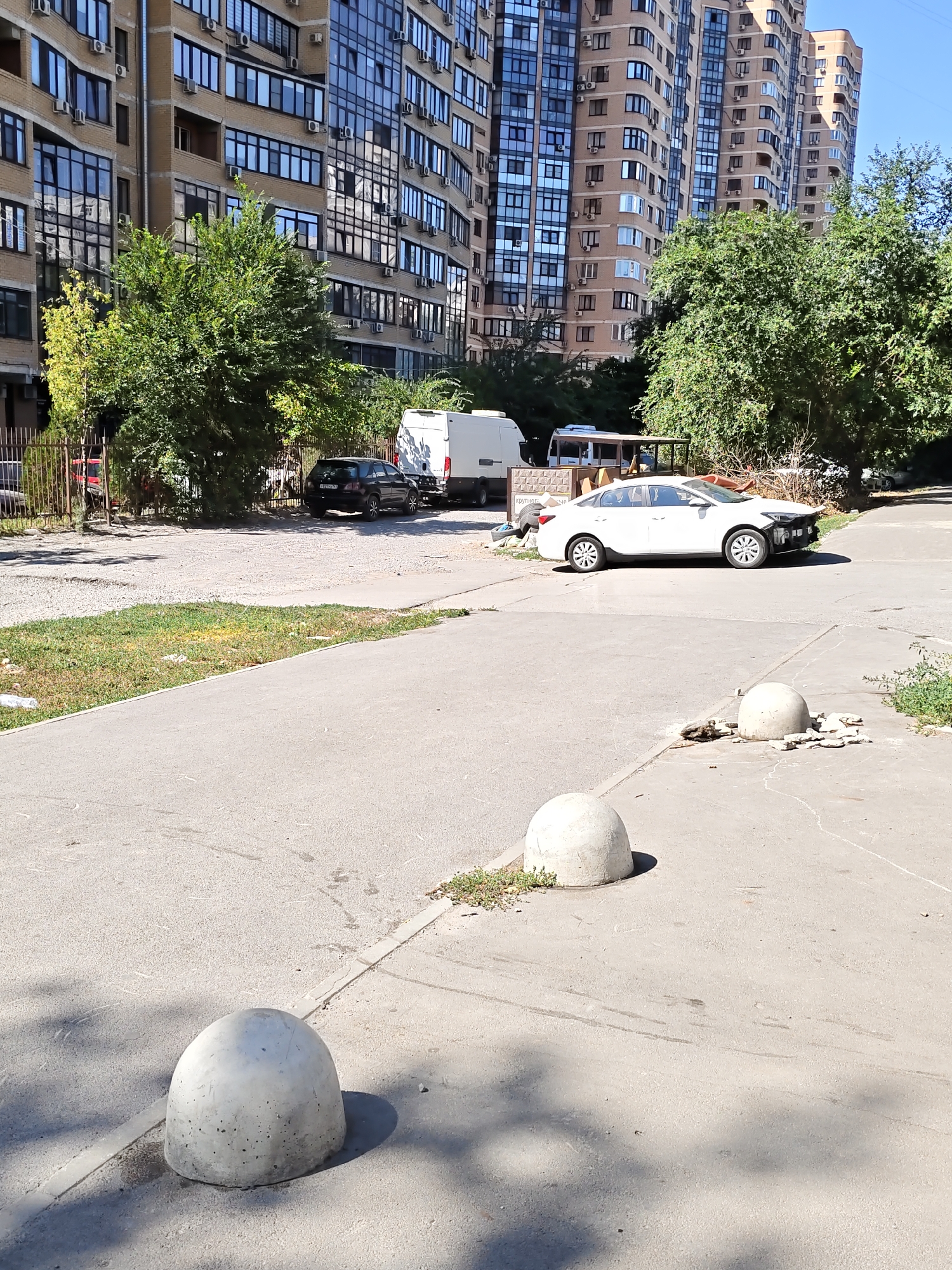 This image depicts an outdoor scene in the city of Rostov-on-Don, Russia, likely a residential courtyard. In the background, there is a large, multi-story apartment building constructed with beige brick and numerous windows, some of which are reflective. Adjacent to the apartment building are several trees. In the middle ground, there is a parking area with several vehicles, including a black car and a white van. A white sedan with visible damage to its front bumper is parked in the foreground. The ground is paved with asphalt and concrete. Several concrete bollards are positioned on the pavement, one of which is in the immediate foreground, and two are in the mid-ground to the right of the white sedan. The sky is clear and blue, indicating a sunny day.