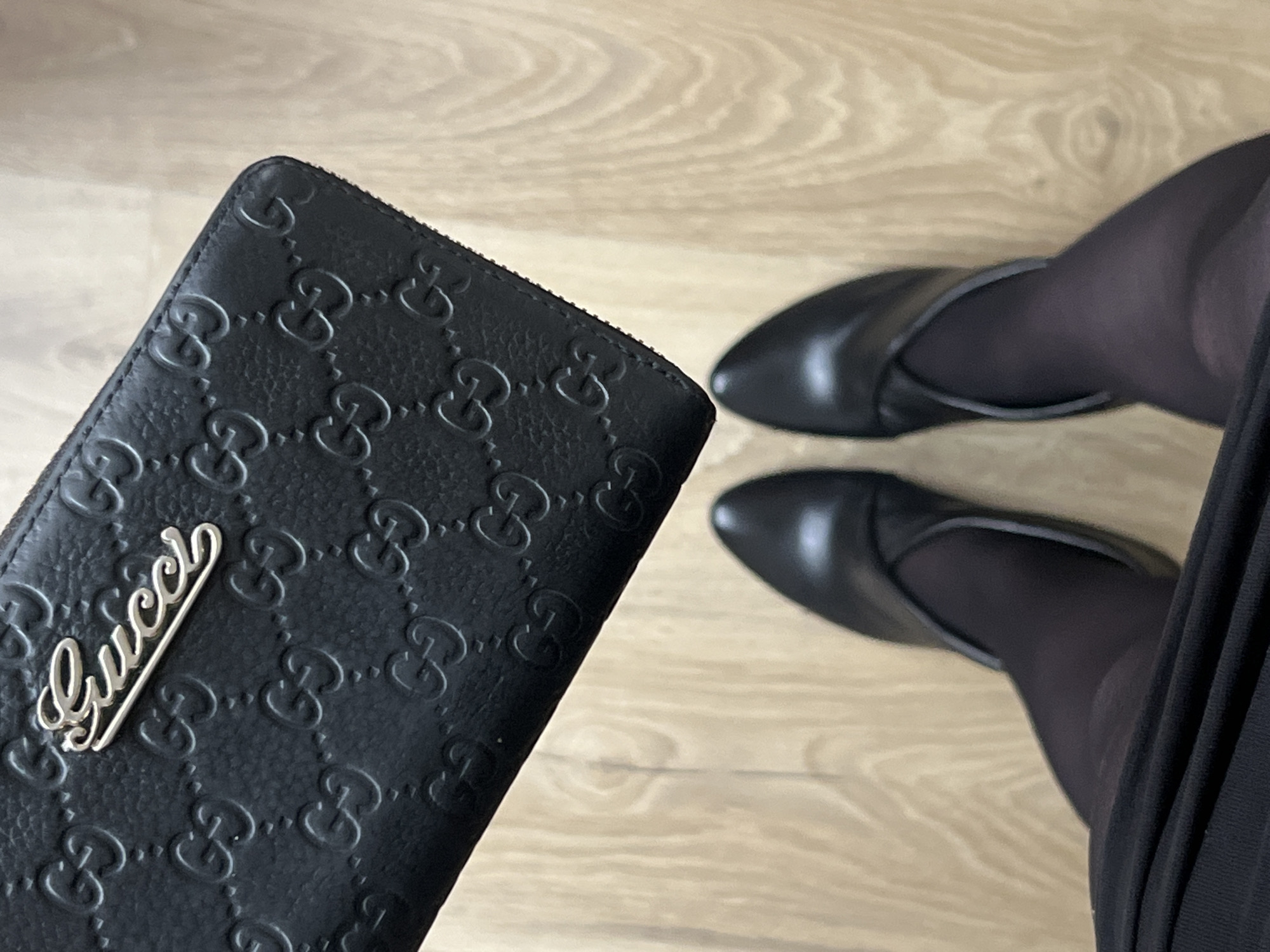 An overhead shot captures a black Gucci zip-around wallet and the lower legs and feet of a person against a light wooden floor. The wallet is made of black textured leather, deeply embossed with the interlocking GG monogram pattern across its surface. A prominent silver-toned metal emblem spelling "Gucci" in a script font is affixed to the front of the wallet.
The person is wearing black, possibly sheer, tights and black pointed-toe flat shoes with a distinctive curved strap detail over the instep. A portion of a dark, possibly pleated, skirt or dress is visible on the right side of the frame. The wooden floor exhibits a subtle grain pattern, indicating an indoor setting. No specific activity is depicted beyond posing with the wallet and feet. The image does not contain any visual cues indicating the time of day, weather, or specific location within Saint Petersburg, Russia, but this is the provided context.