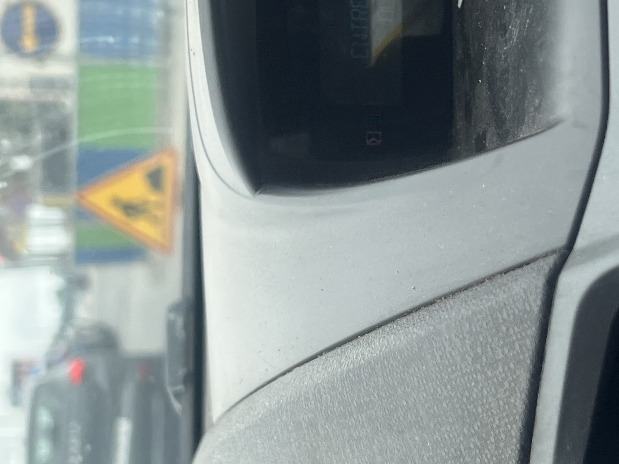 The image captures a perspective from inside a vehicle, looking out through a window while simultaneously showing parts of the car's interior. The exterior scene is significantly blurred, indicating motion or an intentional focus on the vehicle's interior.
Outside the window, several traffic elements are visible through the blur. A blue circular sign with a yellow arrow pointing downwards is in the upper left. Below it, a yellow triangular warning sign with a black symbol, possibly indicating road work or machinery, can be seen. There are also blurry patches of green and blue, possibly from distant signage or landscape. Faint red lights from other vehicles, likely brake lights, are discernible in the bottom left, suggesting traffic.
The vehicle's interior fills the foreground and right side. A textured, gray surface, possibly part of a dashboard or door panel, is prominent in the lower right, transitioning to a smoother, lighter gray area. A dark, recessed screen or display unit is central to the right half of the image. On this display, faint white text reads "ENTREE" (French for "ENTRY"), appearing rotated. Below this, a small red symbol, resembling a charging battery or a phone icon with a lightning bolt, is visible, followed by the numeral "3" in red.
The scene appears to take place during the daytime. The perspective suggests someone is in the passenger or driver's seat of a moving vehicle. The visible French text "ENTREE" aligns with the provided location context of Casablanca, Morocco.