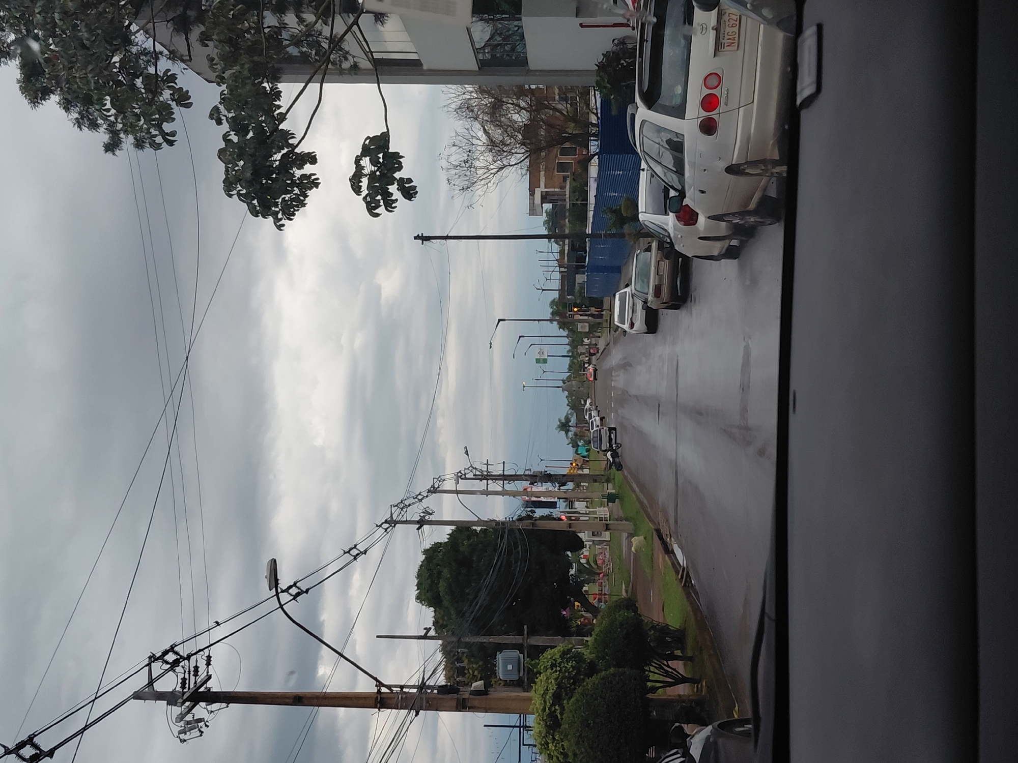 The image shows a street scene in Encarnacion, Paraguay, on a cloudy day. The perspective is from inside a vehicle, looking forward. A wet asphalt road is lined with cars, trees, and utility poles with many wires. On the right side of the street, there is a modern building with windows. On the left side of the street, there are more trees and some manicured bushes. In the distance, there are more cars and buildings, and the sky is filled with heavy clouds. The overall impression is a somewhat grey and overcast urban environment.