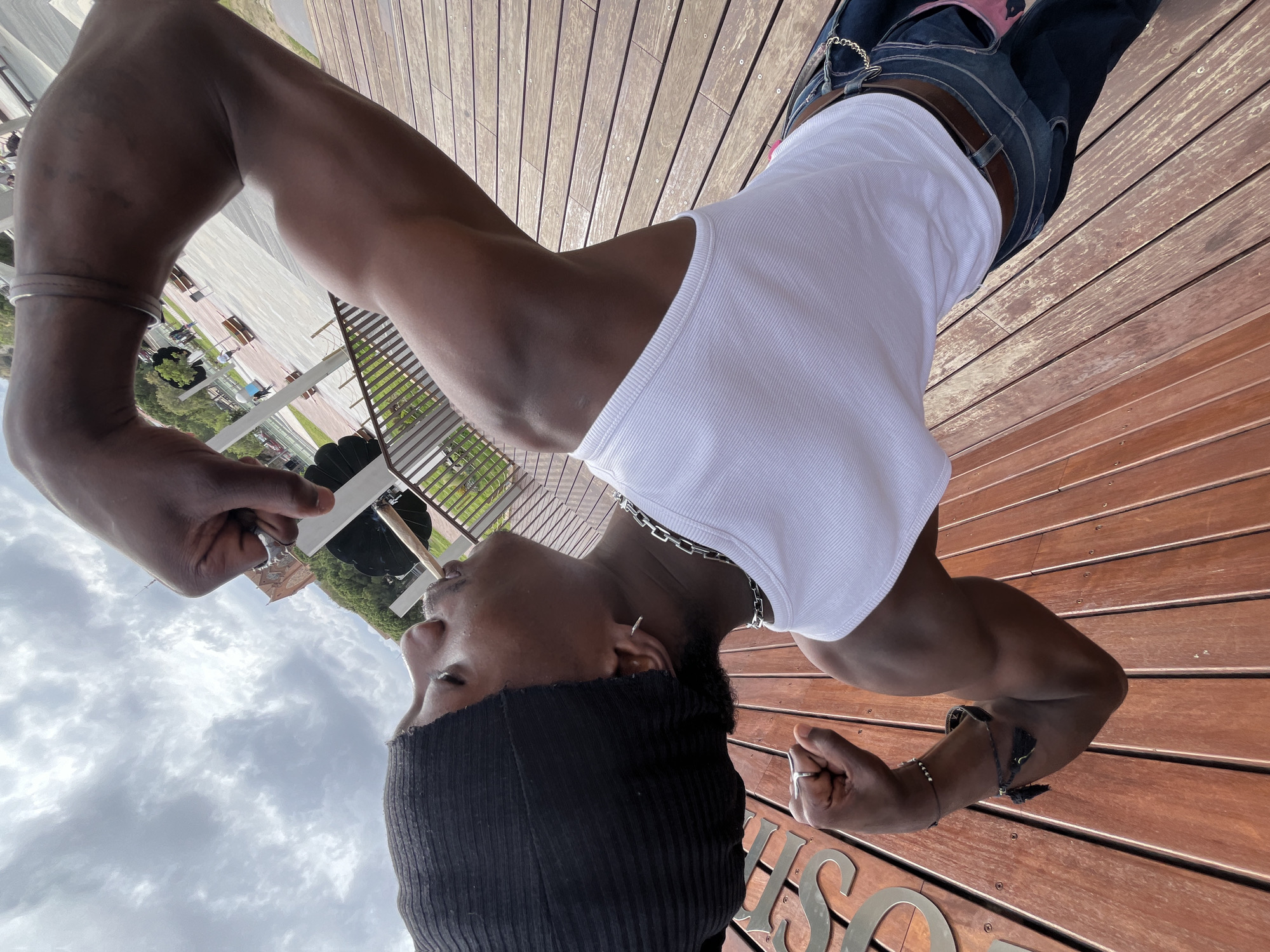 A medium-shot, upside-down image depicts a person of color standing on a wooden platform, facing the sky. The person is wearing a black beanie, a white ribbed tank top, and denim pants with a brown belt. They are adorned with silver jewelry, including bracelets, rings, and a chain necklace. The background includes a partially visible urban park or garden area with greenery and benches. The presence of wooden planks and signage suggests the image was taken in a city. Based on the visible objects and the urban setting, the location is most likely in Barcelona, Spain, though further specific landmarks would be needed to pinpoint the exact location.