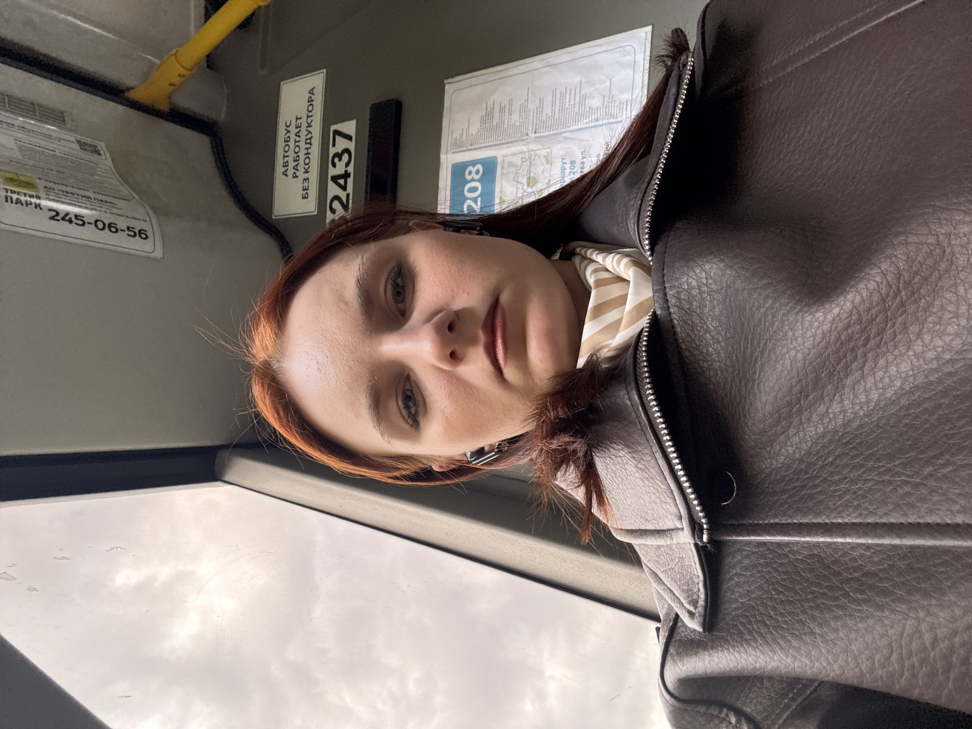 A woman is taking a selfie on a bus in Saint Petersburg, Russia. The bus has a sign indicating it is route 208 and operated by "Third Park" (Трети парк), with the phone number 245-06-56. There is also a notice stating "Автобус работает без кондуктора" (Bus operates without a conductor) and the bus number 2437. The woman is wearing a brown leather jacket and a striped scarf. The bus window is visible, with a cloudy sky outside.